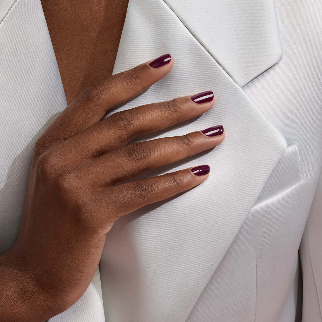 A hand with Prune, a purple vintage nail polish by Manucurist, rests on the lapel of a white suit jacket, highlighting the neat manicure and smooth, tailored fabric.