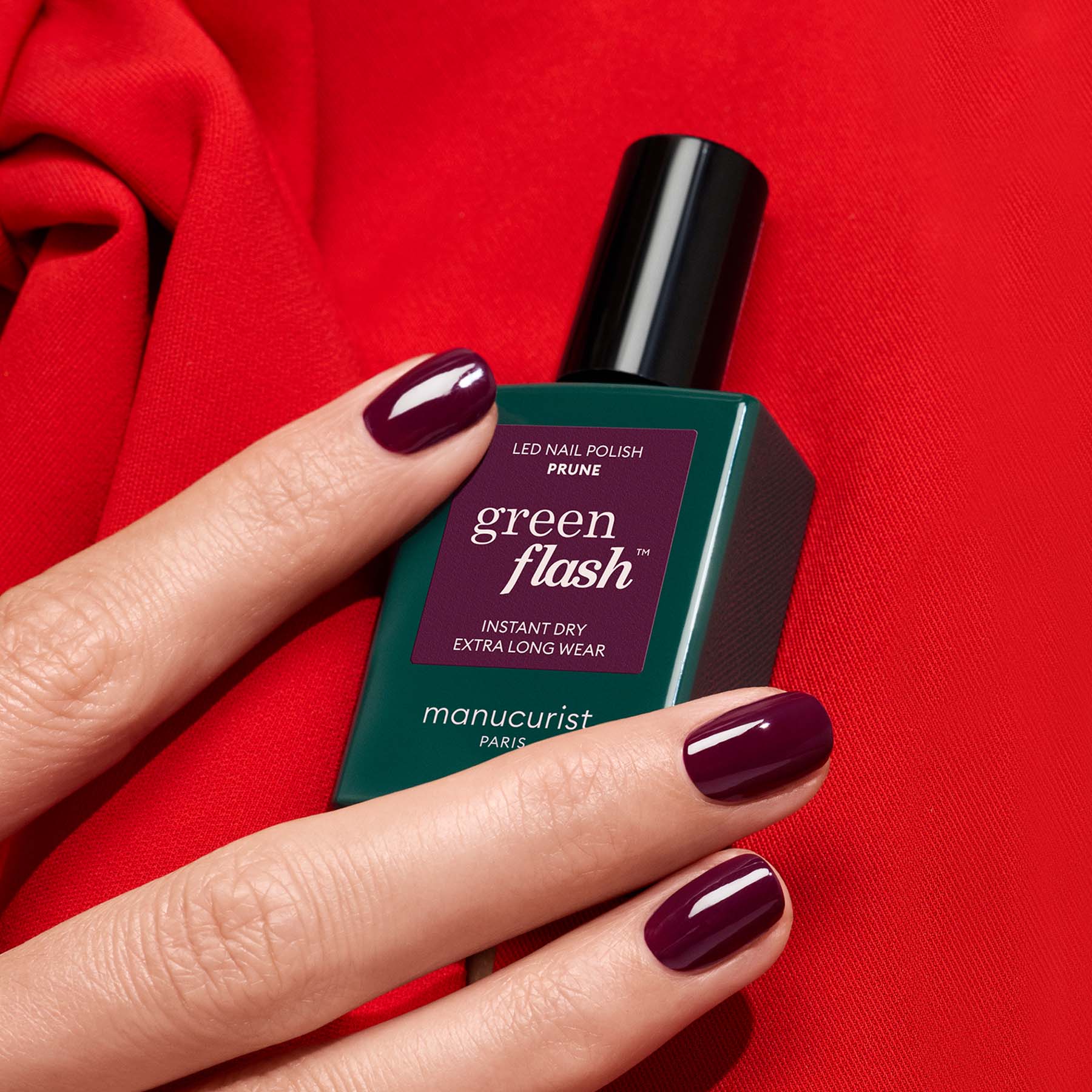 A hand with deep wine red gel polish holds a bottle of Manucurist Prune nail polish against a bright red fabric background.