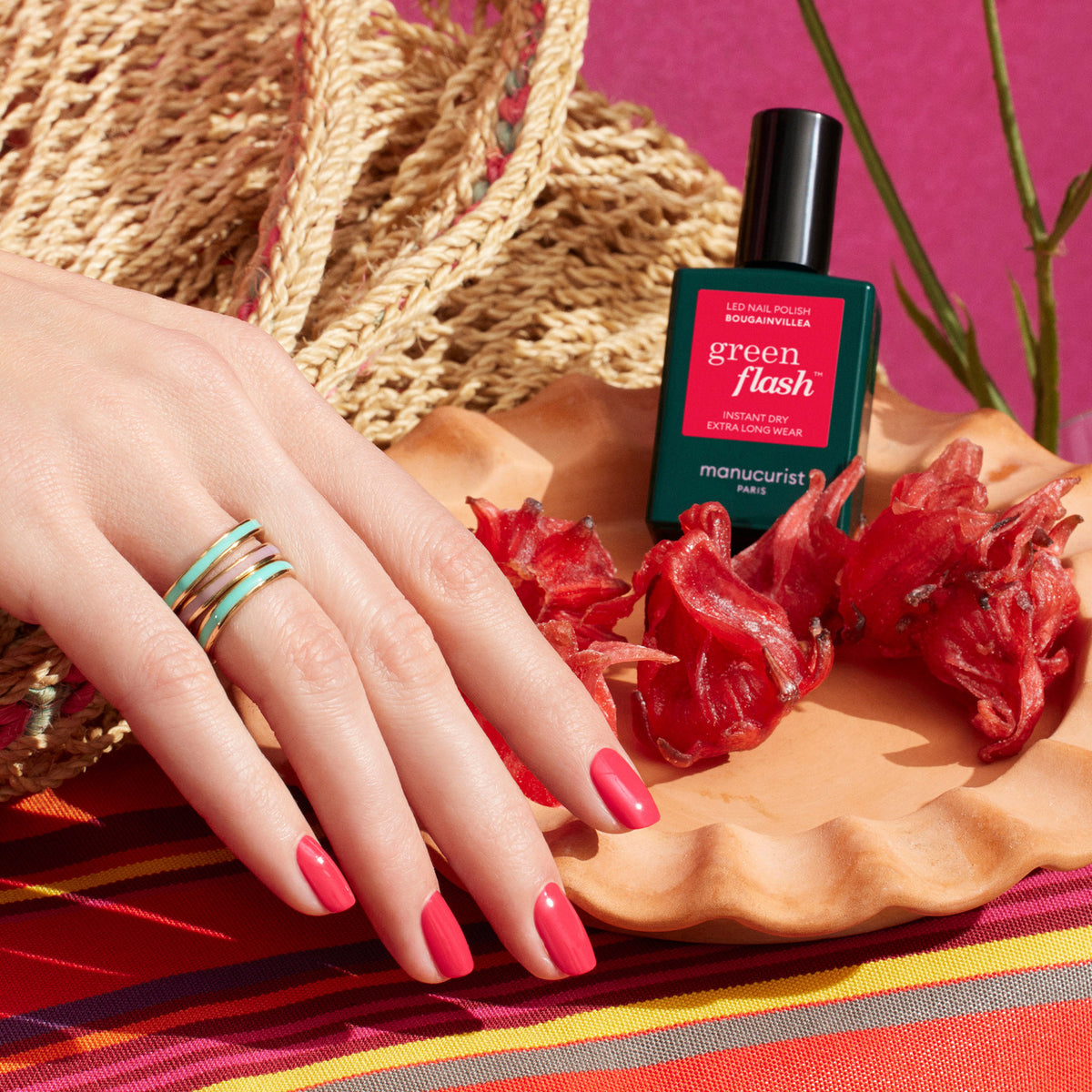 A hand with bright red Manucurist Bougainvillea polish rests on a clay dish with dried bougainvillea, next to the gel bottle, against a backdrop of a woven bag and a colorful striped cloth.