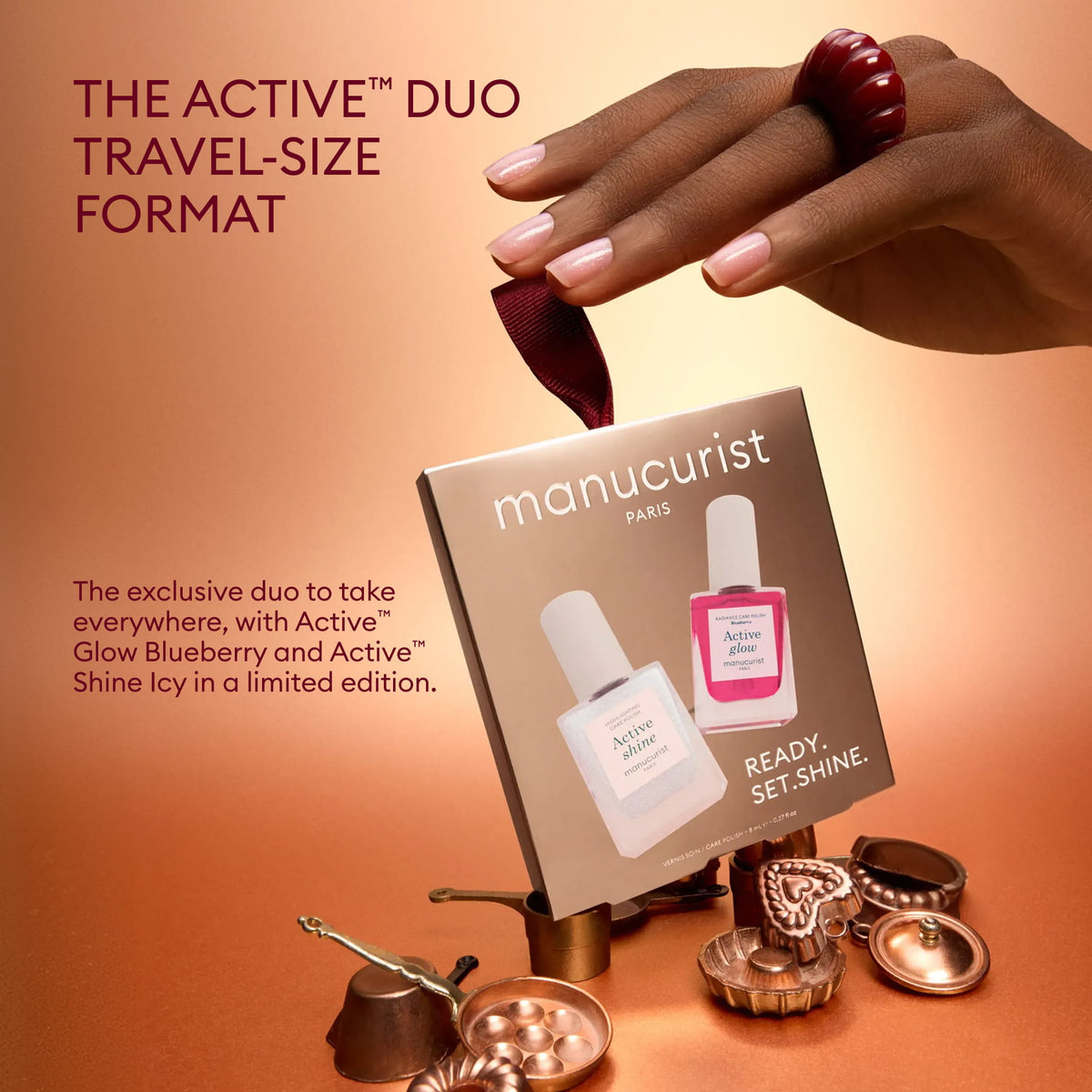 A manicured hand holds a ribbon attached to a box containing two bottles of Manucurist’s Active™ Travel-Size Duo, surrounded by metallic objects, with text highlighting it as the perfect holiday gift set for nail care enthusiasts.