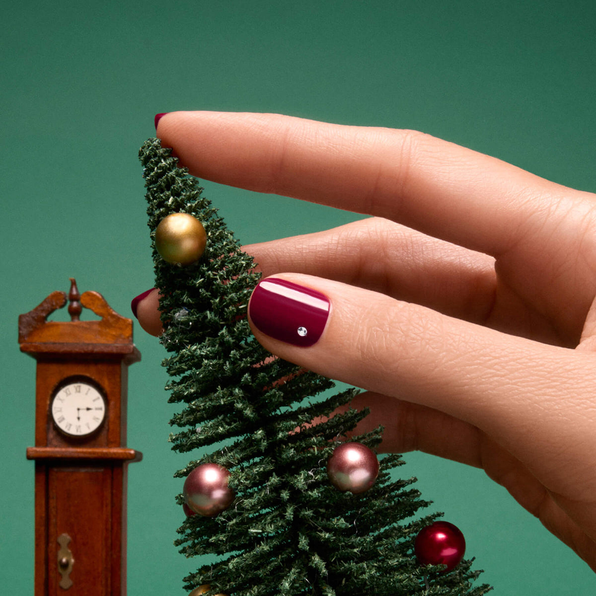 A hand with a holiday manicure featuring burgundy polished nails decorates a small artificial Christmas tree adorned with ornaments, next to a miniature grandfather clock, set against a green background. Sparkling accents from Manucurist Rhinestones Nail Art Holidays add festive charm to the look.