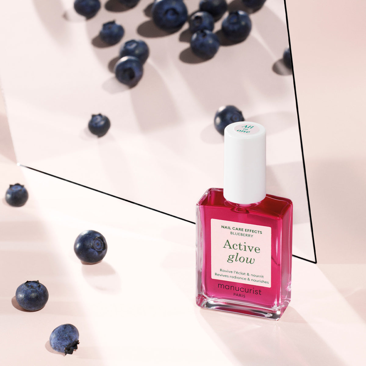 A bottle of Manucurist’s Active™ Glow Blueberry nail polish sits on a pale pink surface with scattered blueberries and its reflection visible in a mirror behind it, offering radiant shine and nourishing nail care for a flawless look.