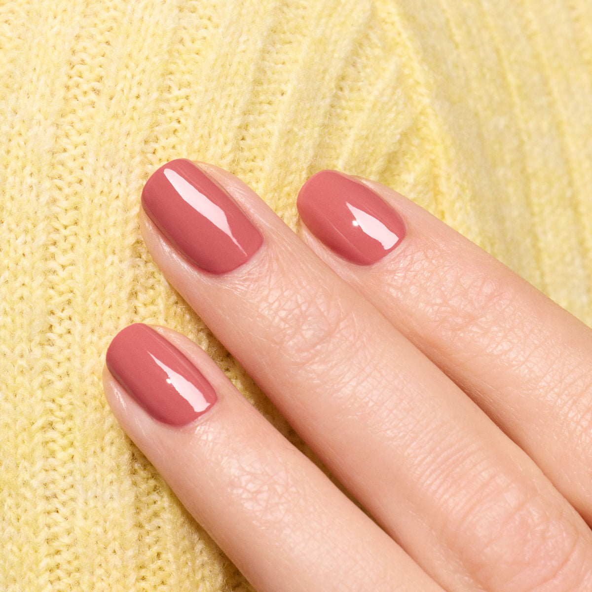 A close-up of a hand with neatly manicured nails painted in glossy Bois de Rose polish from Manucurist’s Green™ range, resting on textured yellow knitted fabric.