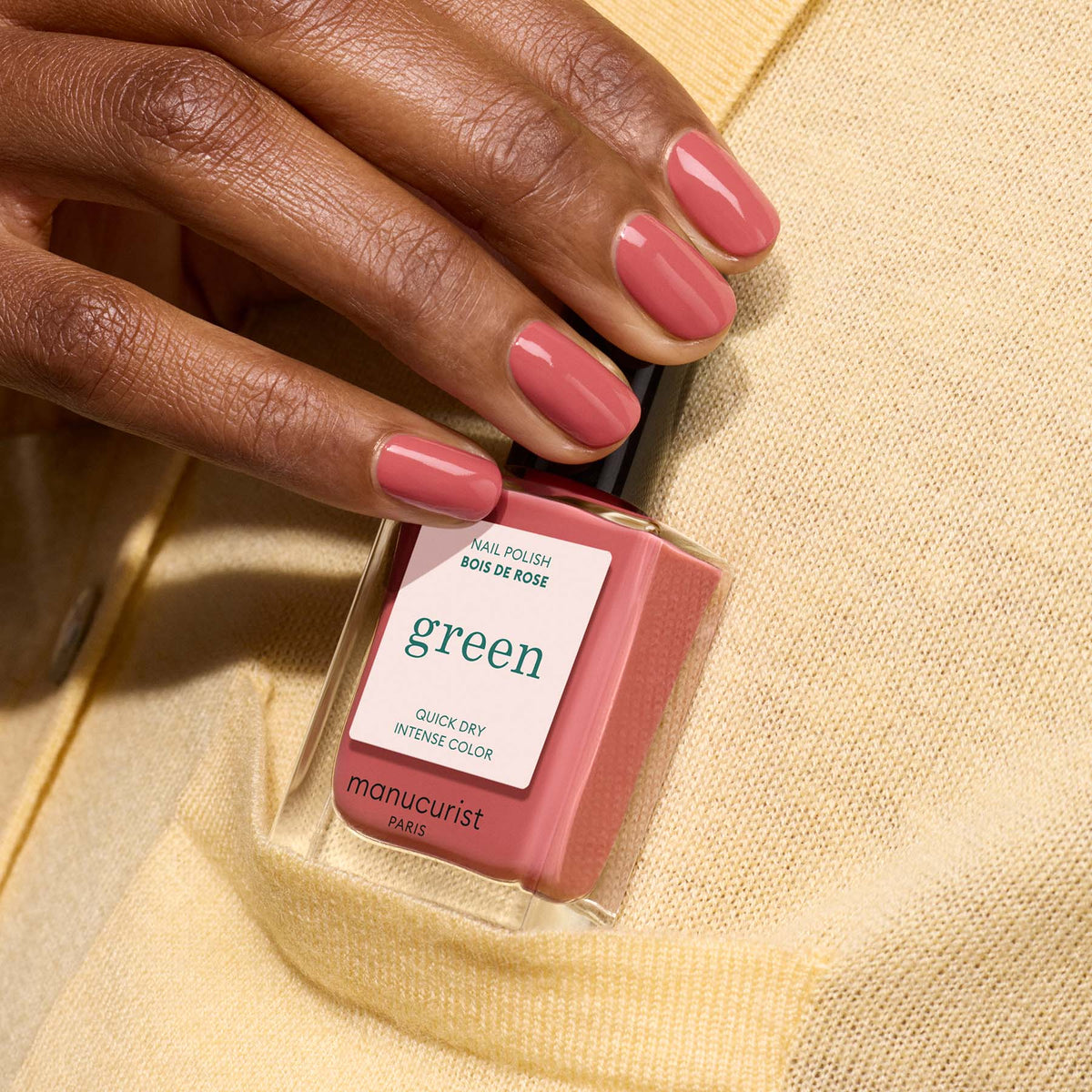 A hand with neatly manicured nails in Bois de Rose holds a Manucurist nail polish bottle labeled green, set against a beige sweater.
