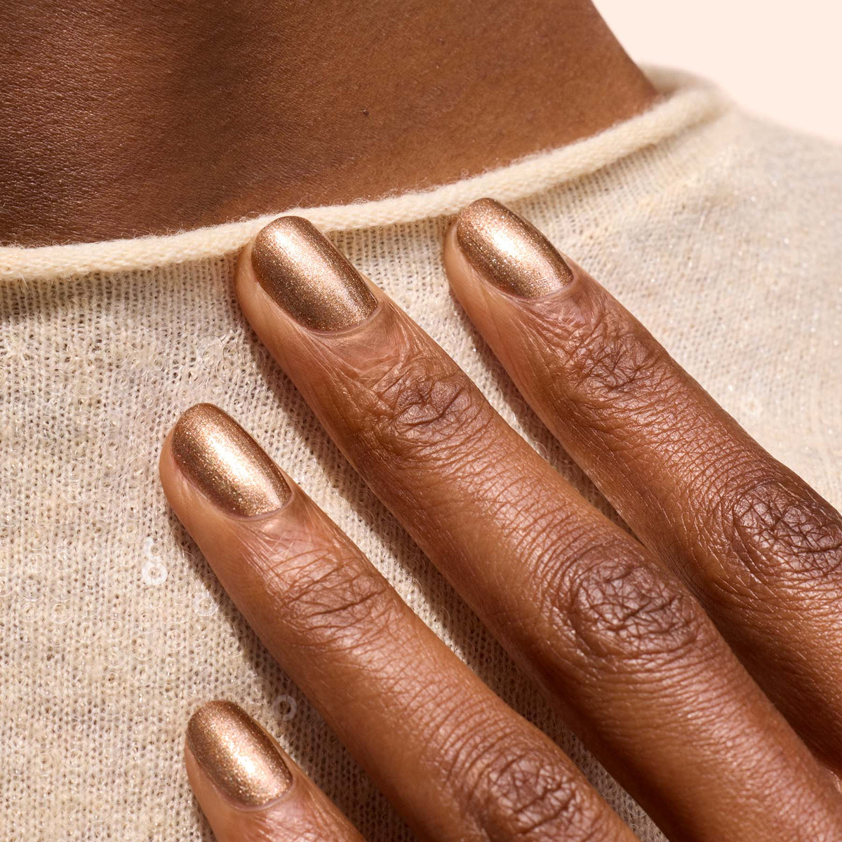 A close-up of neatly shaped nails painted with Manucurist’s Bronzé brown glitter polish, resting on deep brown skin against a light textured sweater.