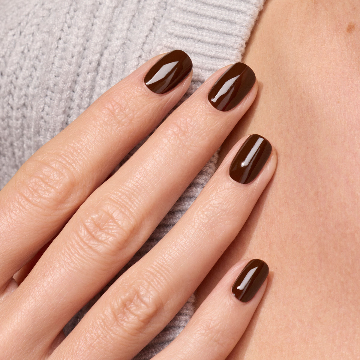A hand with neatly manicured nails painted in Manucurist’s Clove, a glossy chocolate brown shade, rests against a light gray ribbed sweater.