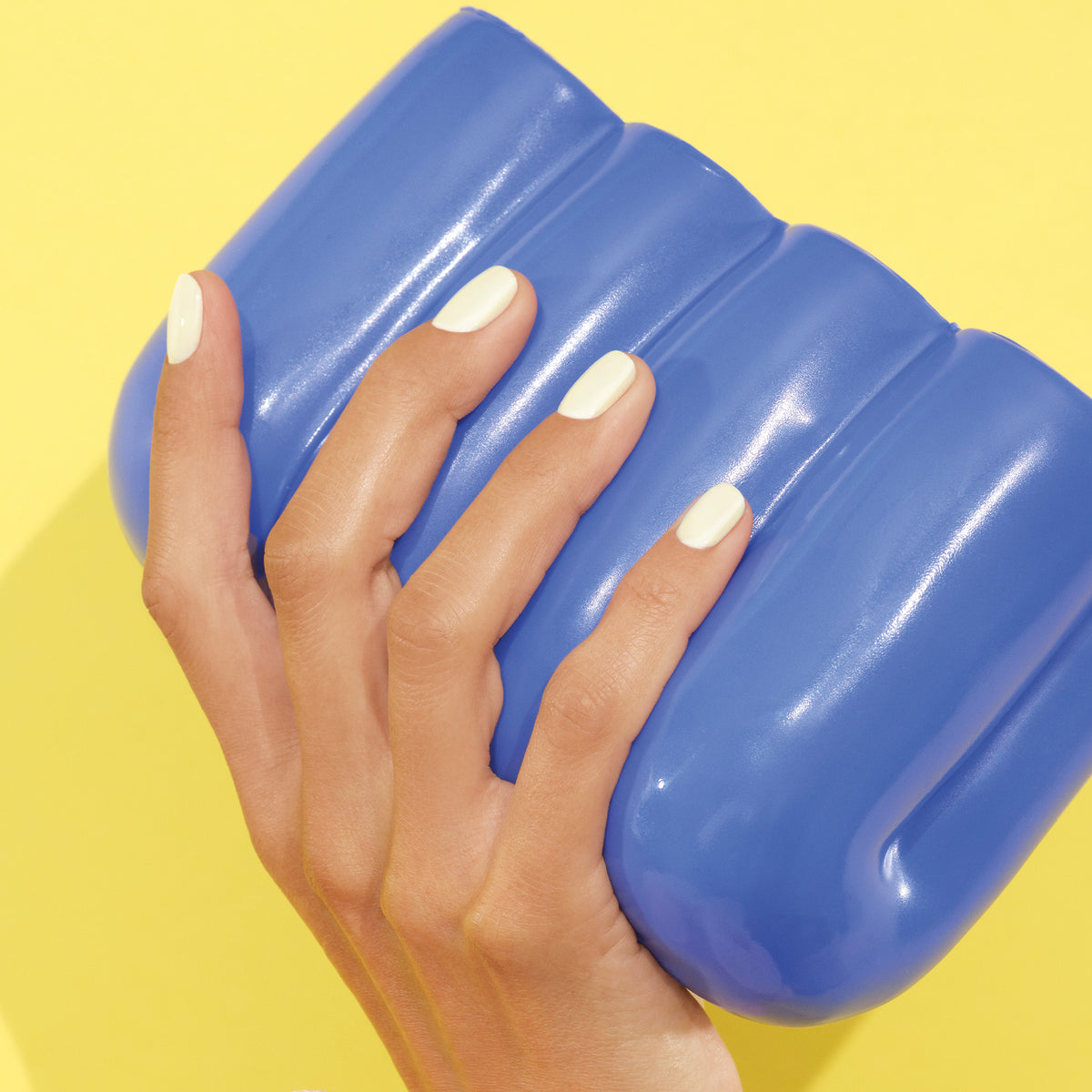 A hand with neatly manicured Porcelaine gel polish nails by Manucurist holds a large, shiny blue object against a bright yellow background.