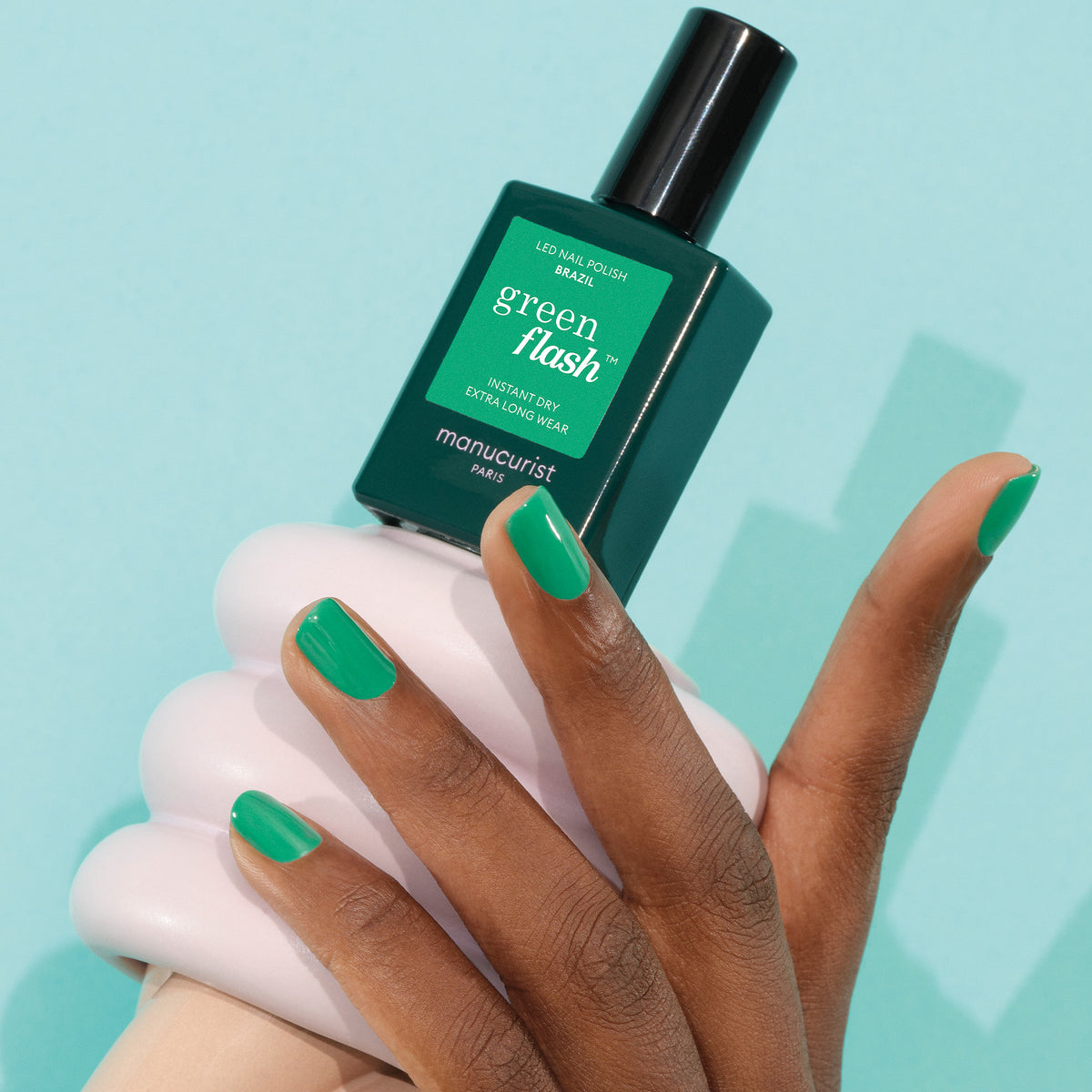A hand with medium brown skin tone displays glossy electric green nails, holding a bottle of Manucurist’s Brazil polish against a light blue backdrop.