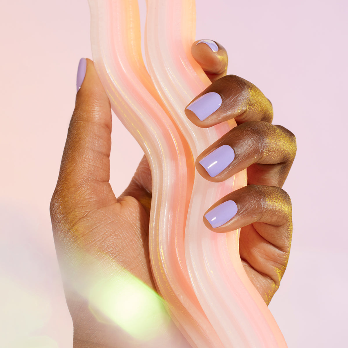 A hand with Manucurist’s Wisteria pastel purple gel polish holds a wavy, pastel swirl object, set against a soft light pink background.