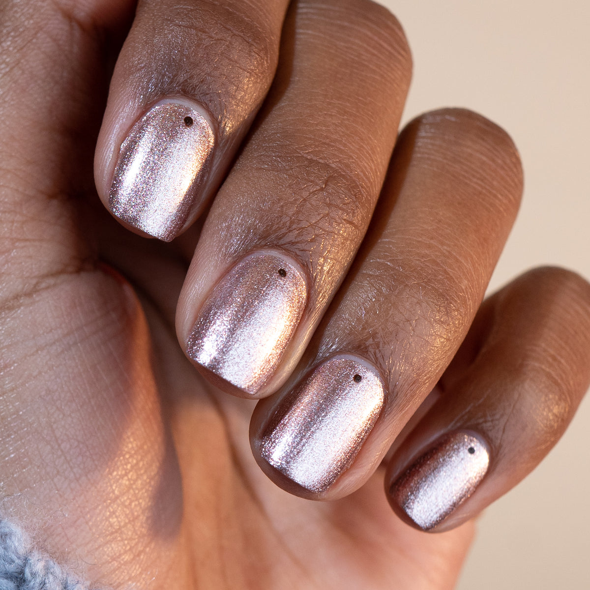 A close-up of a hand with brown skin displays four fingernails painted in Manucurist’s Green™ polish, Cosmic Rose—an iridescent pink shade—each nail adorned with a small black dot near the cuticle.