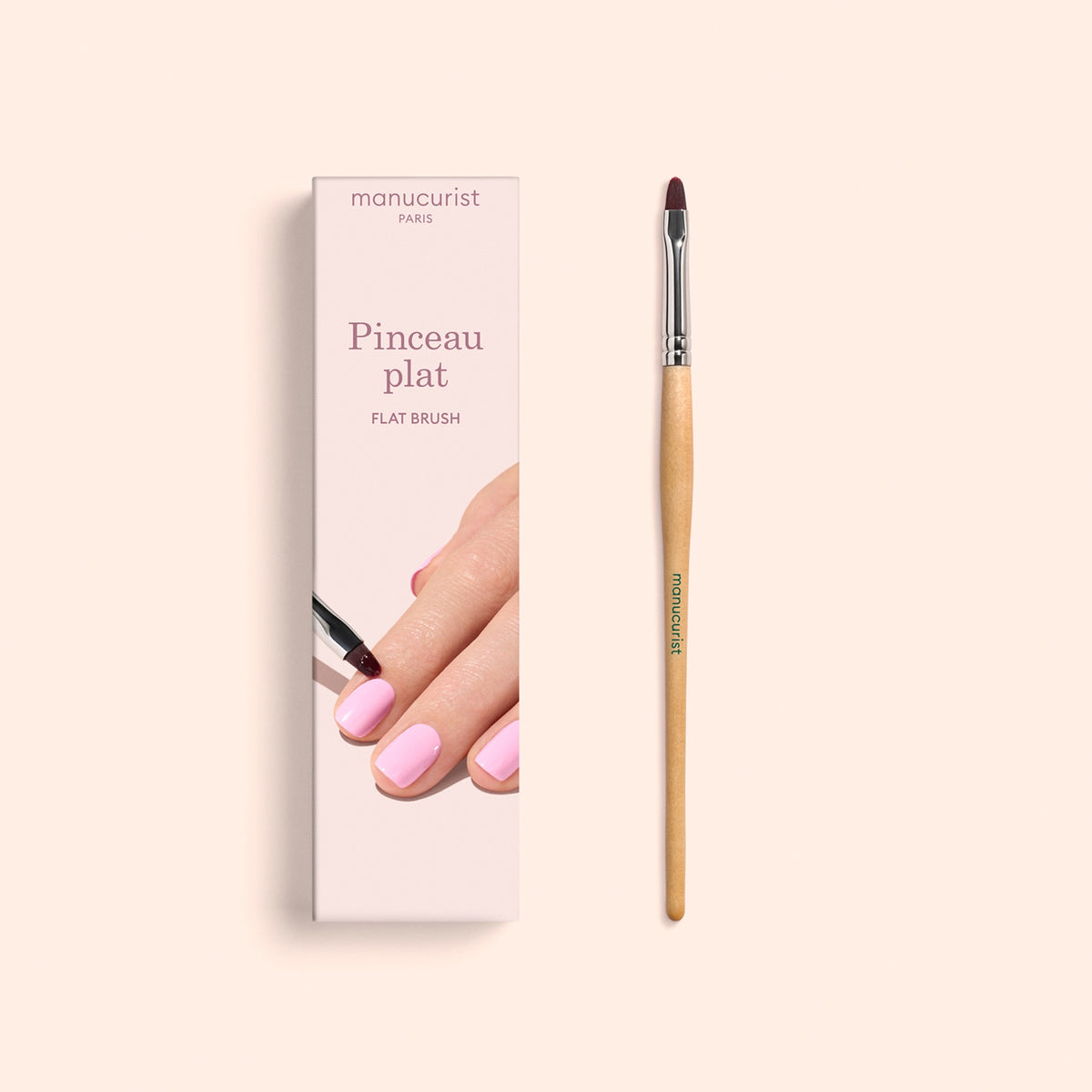 A Manucurist Flat Brush with a wooden handle lies next to its box, which features an illustration of a hand with pink-painted nails along with the text “Pinceau plat” and “Flat Brush”—ideal for precise manicure application and cuticle care.
