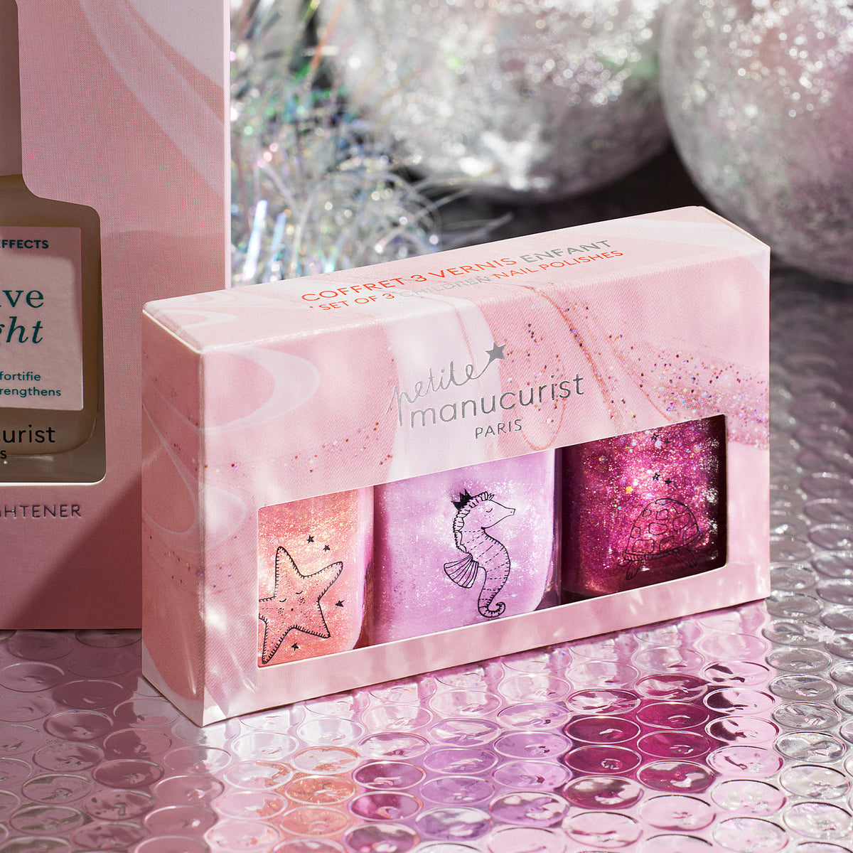 The Petite Manucurist Trio by Manucurist features three sparkly nail polishes in a pastel pink box with starfish, seahorse, and shell designs, displayed on a shiny textured surface with glittery decorations in the background.