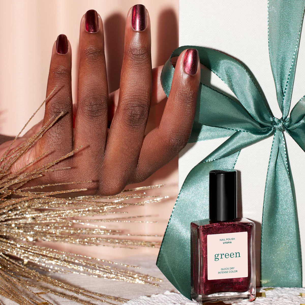 A hand with burgundy polish rests near a white gift box tied with a green ribbon. In front is Manucurist’s Utopia, a metallic crimson nail polish, while decorative golden branches complete the scene.
