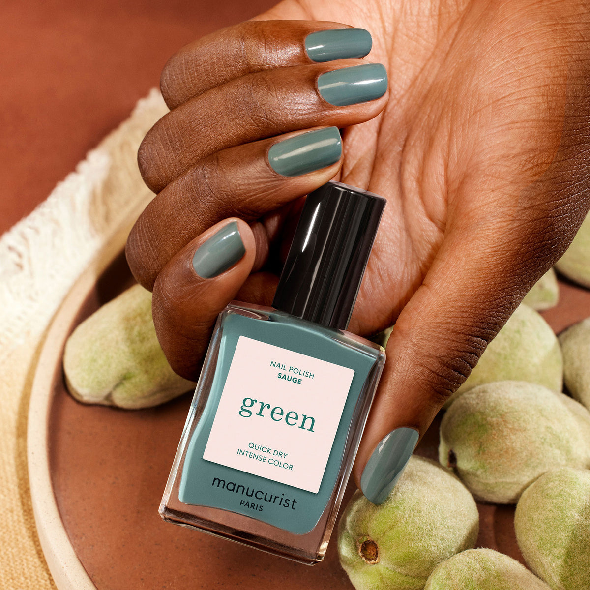 A hand with teal-green painted nails holds a matching bottle of Manucurist Sage nail polish. Unripe almonds are arranged on a brown surface in the background.