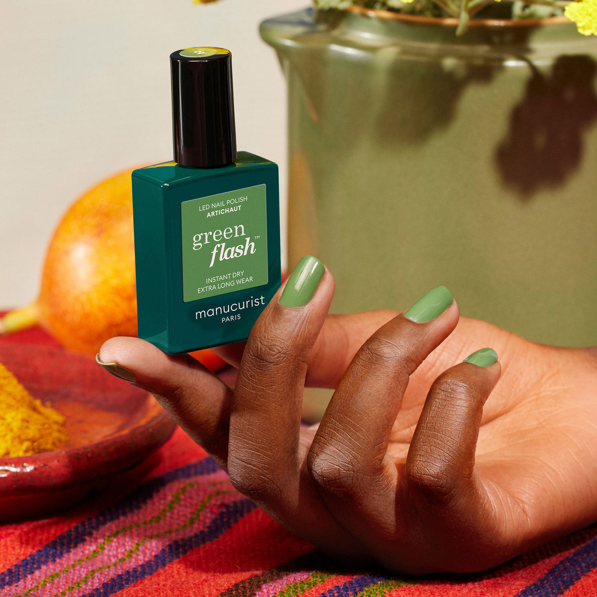 A hand with Artichoke green nail polish holds a matching Manucurist bottle. In the background are a patterned cloth, an orange, and a green vase with flowers.