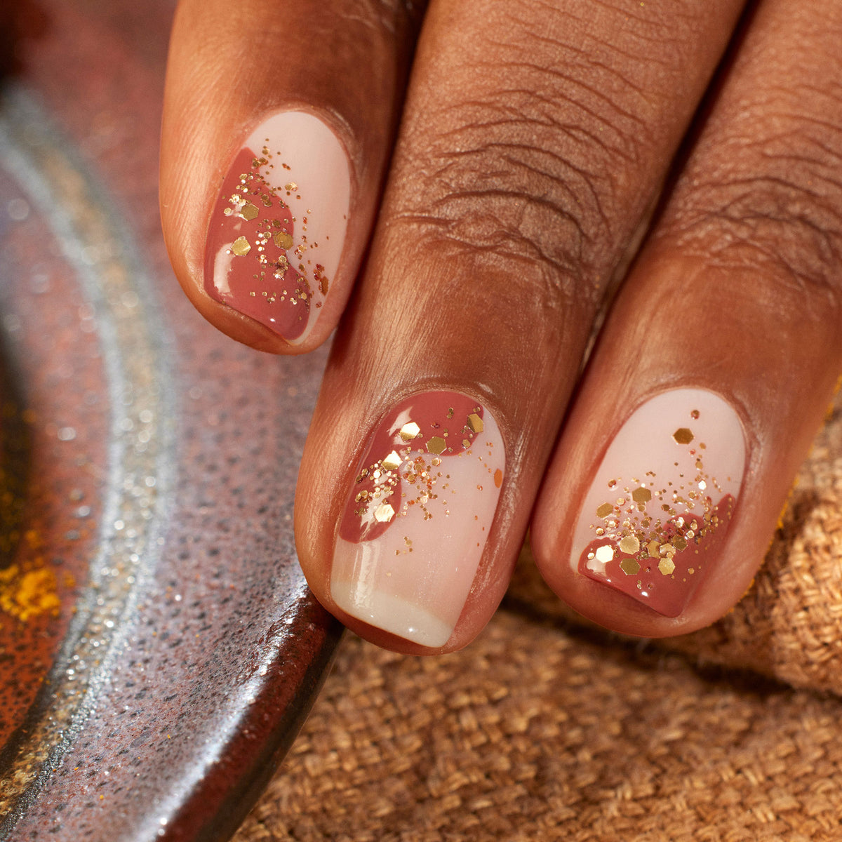 Close-up of a hand with natural nails in a pink and nude ombre, adorned with gold glitter and brick red Brique by Manucurist. The medium brown skin tone contrasts textured brown and metallic backgrounds.