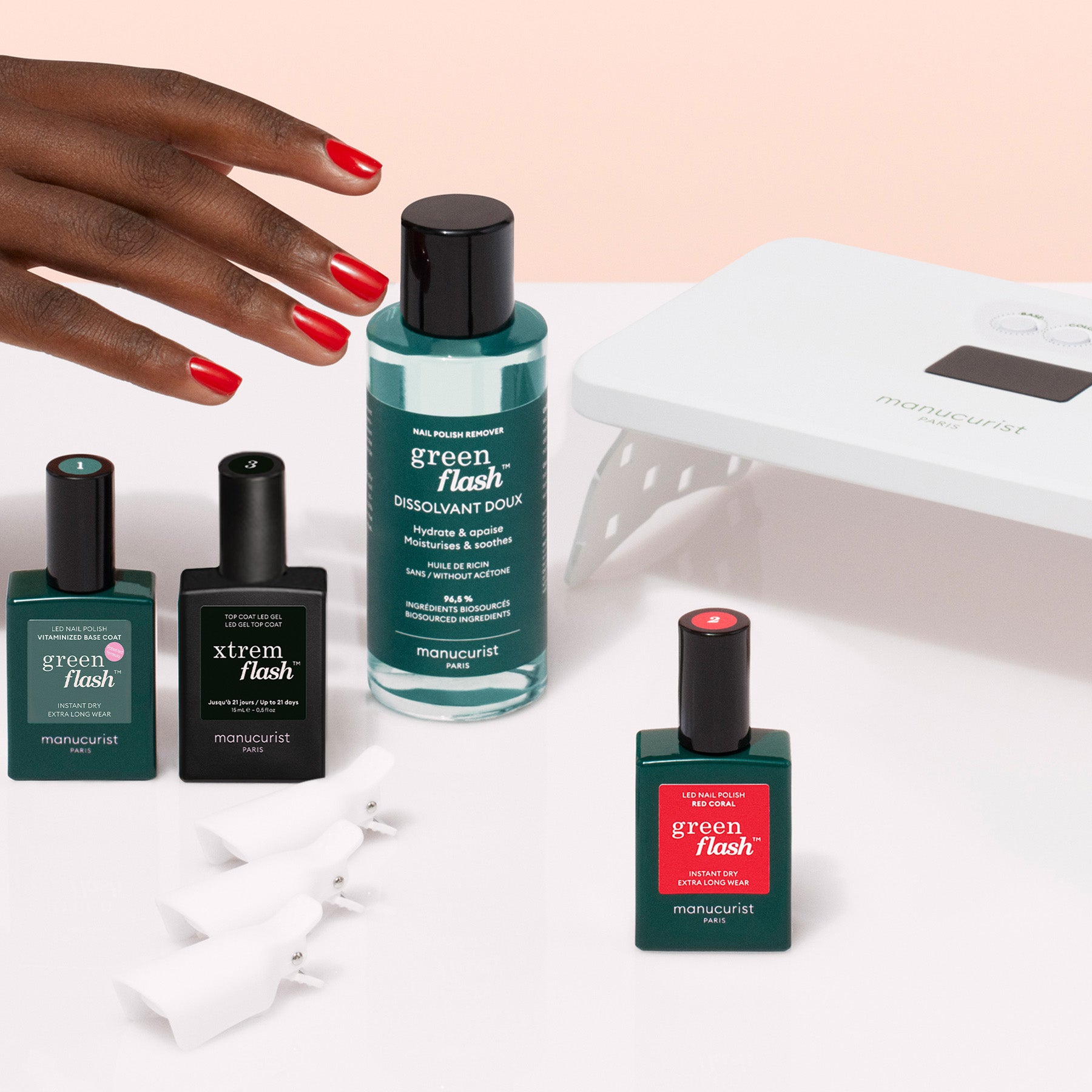 A hand with red nails reaches for the Manucurist Discovery Kit, which includes gel polish bottles in green, black, and red with labeled caps, a nail lamp, and white nail clips for a long-lasting manicure.