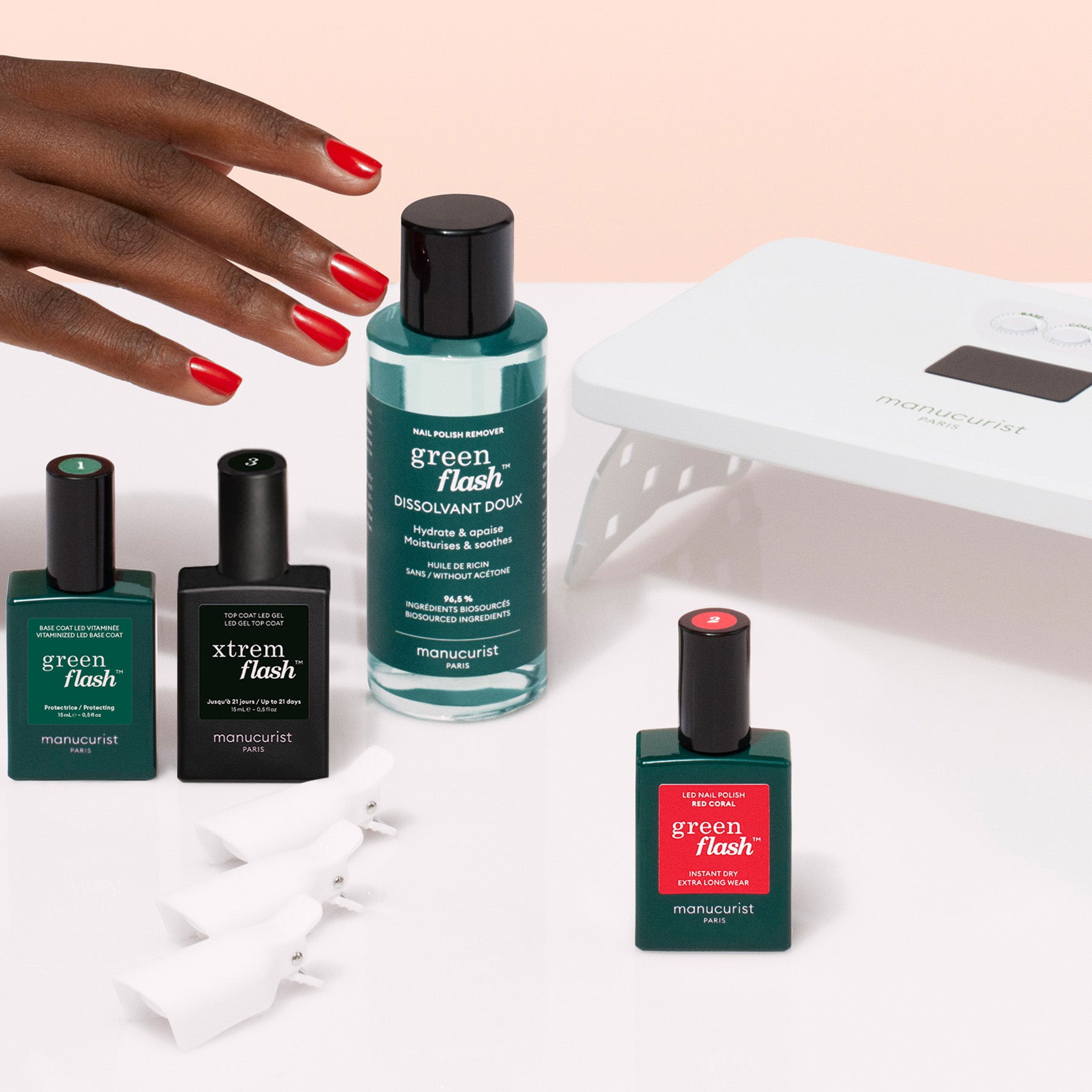 A hand with red nails reaches for the Manucurist Discovery Kit, which includes gel polish bottles in green, black, and red with labeled caps, a nail lamp, and white nail clips for a long-lasting manicure.