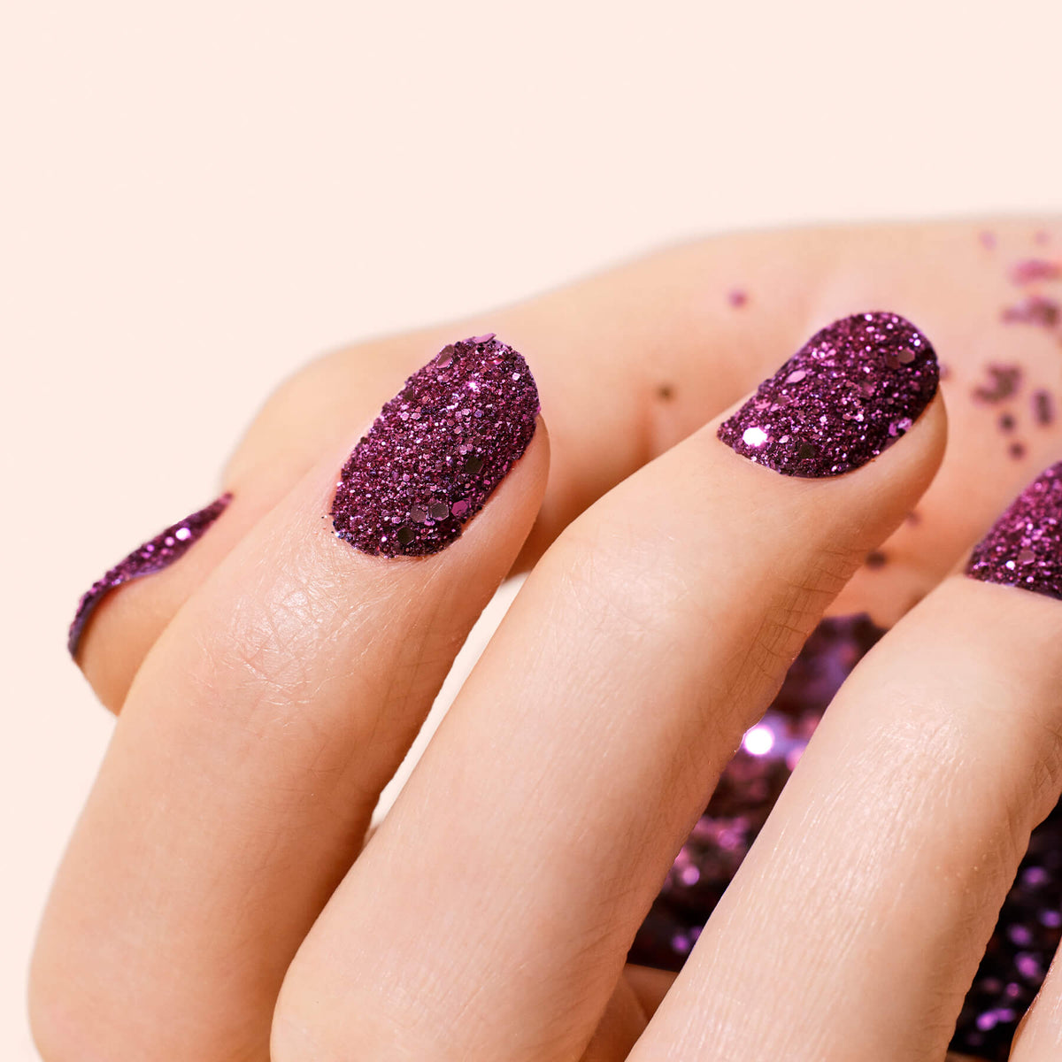 A close-up of a hand with sparkly purple gel polish and Manucurist Bordeaux biodegradable glitter scattered on the skin and background.