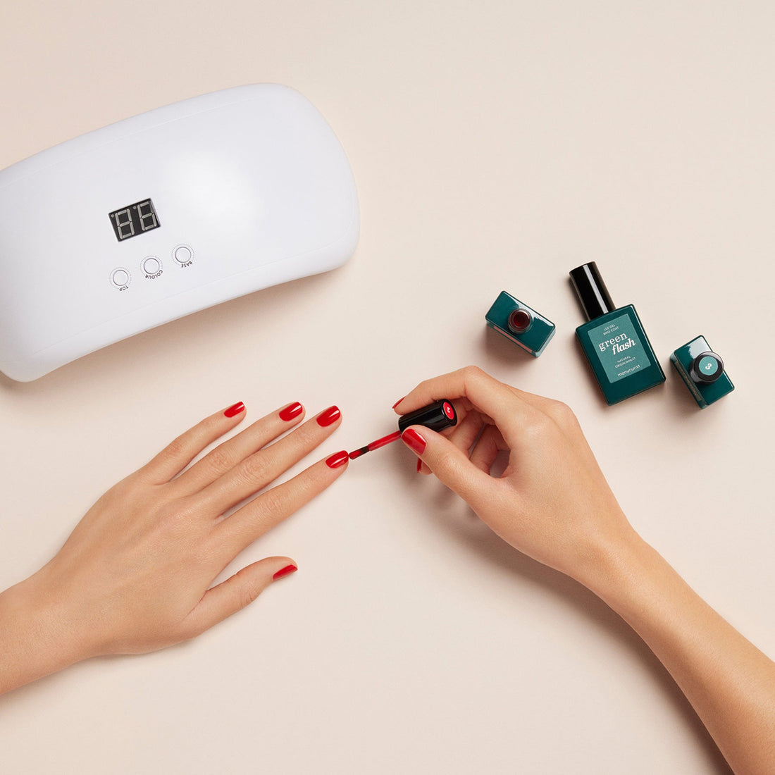 A person applies red polish from the Manucurist Welcome Kit Green Flash™ to their nails. Teal bottles and a white LED nail lamp are nearby on a beige surface.