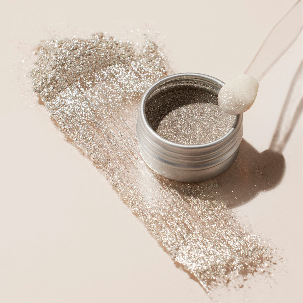 The Manucurist Glitter Duo Silver Mirror loose powder jar, complete with a foam applicator, sits on a light surface. A spill from the open jar creates a sparkling streak of pearlescent shine beside it.
