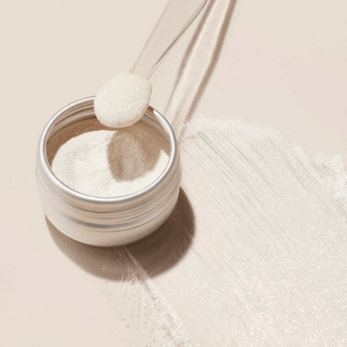 A small metal container of Manucurist Glitter Duo Rose Gold Glazed holds fine white powder, with a clear plastic spoon on top and some powder spread over a pale surface. The scene is simple and softly lit.