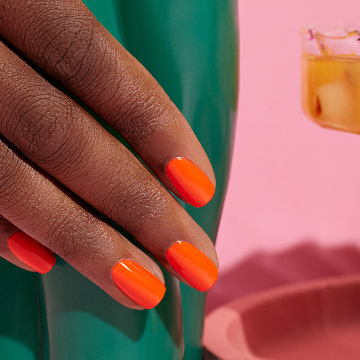 A hand with smooth brown skin and glossy orange nails painted with Manucurist Flower love rests on a green surface. The background is pink, with a blurred cup holding an iced drink off to the side.