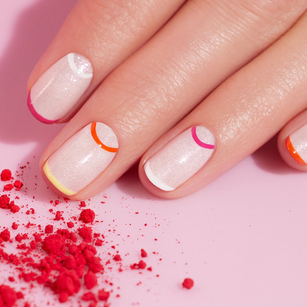 A hand with shimmery pale pink Smile! nail polish from Manucurist features colorful half-moon designs made using a Nail Art Liner Brush, resting on a light pink surface—perfect summer nail art inspiration.