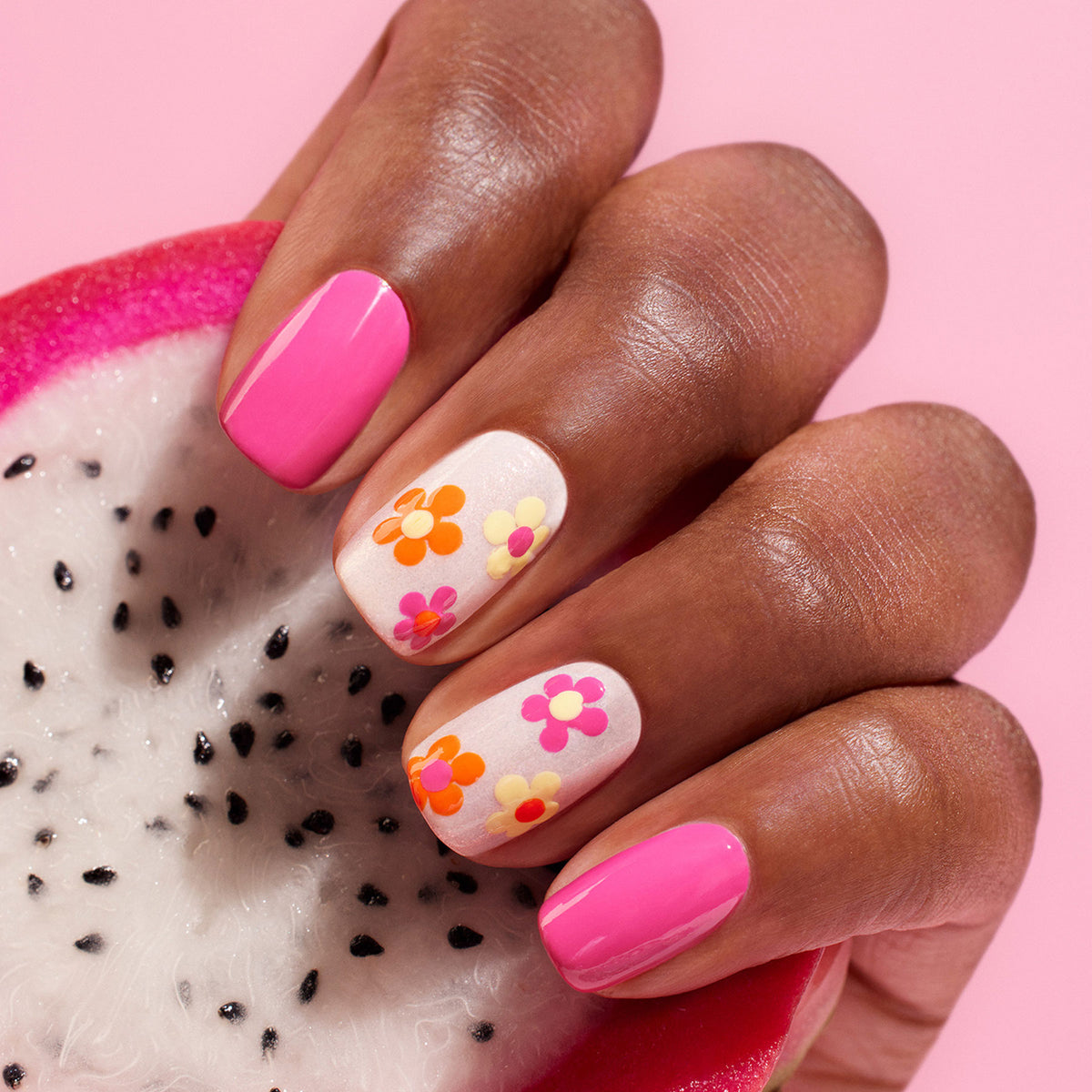 A hand with pink and white "Flower love" nails by Manucurist, adorned with colorful spring flower designs, holds a sliced dragon fruit against a pink background.