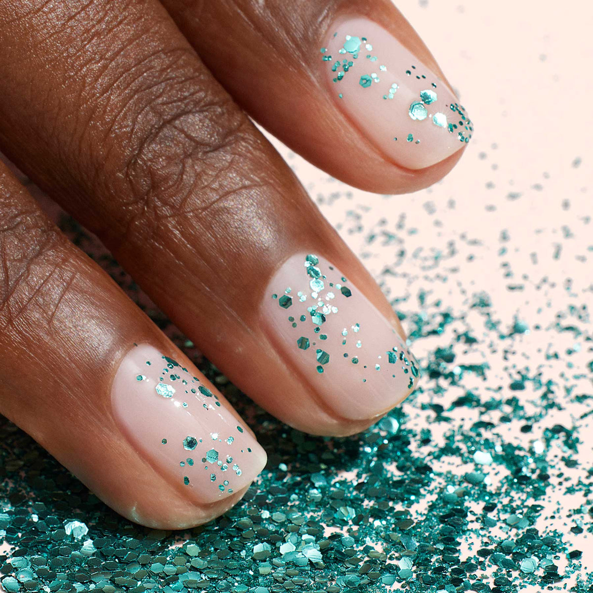 A close-up of natural nails painted with clear polish and decorated using Manucurist Turquoise biodegradable glitter, resting on a surface sprinkled with the same eco-friendly turquoise glitter.