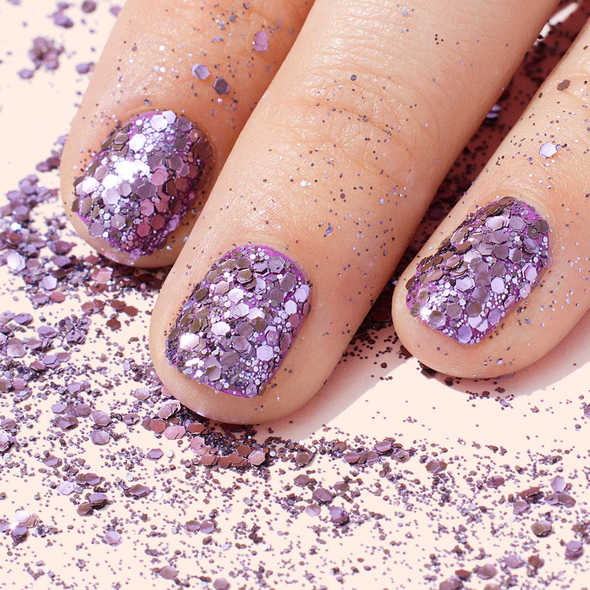 Close-up of fingers with nails painted in Manucurist Lilac biodegradable glitter polish, surrounded by scattered Manucurist Lilac biodegradable glitter on a light surface.