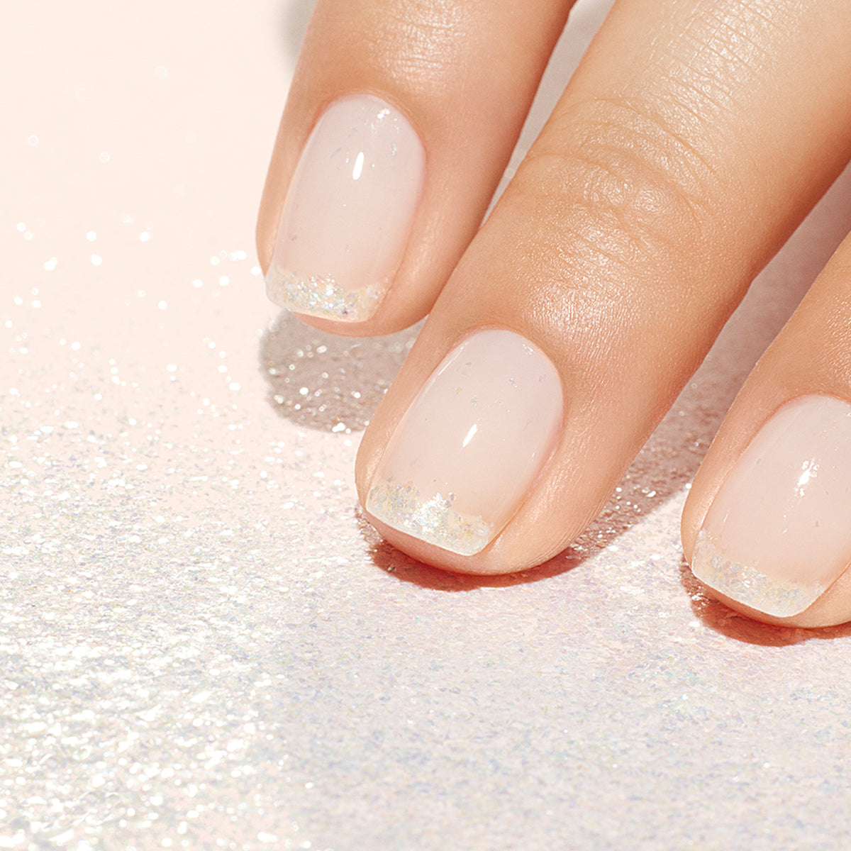 A close-up of a hand with a Manucurist Green Flash manicure, featuring nails painted with Nacres Flakes—a natural pink base and glittery silver tips with pearlescent accents—resting on a sparkly, light-colored surface.