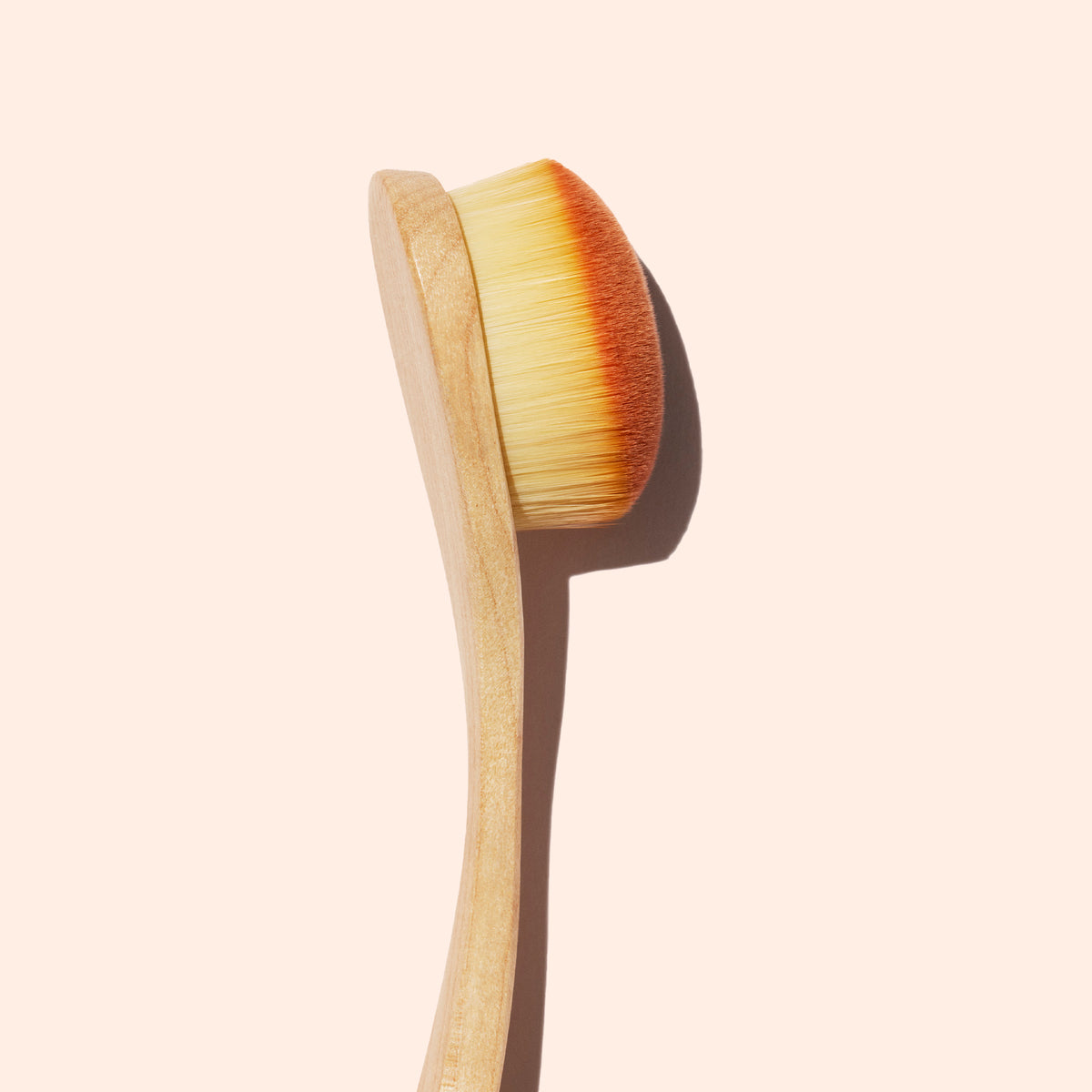 A close-up of the Manucurist Dust Brush, featuring a wooden handle and dense, soft vegan bristles, set against a plain beige background with a shadow cast to the right.