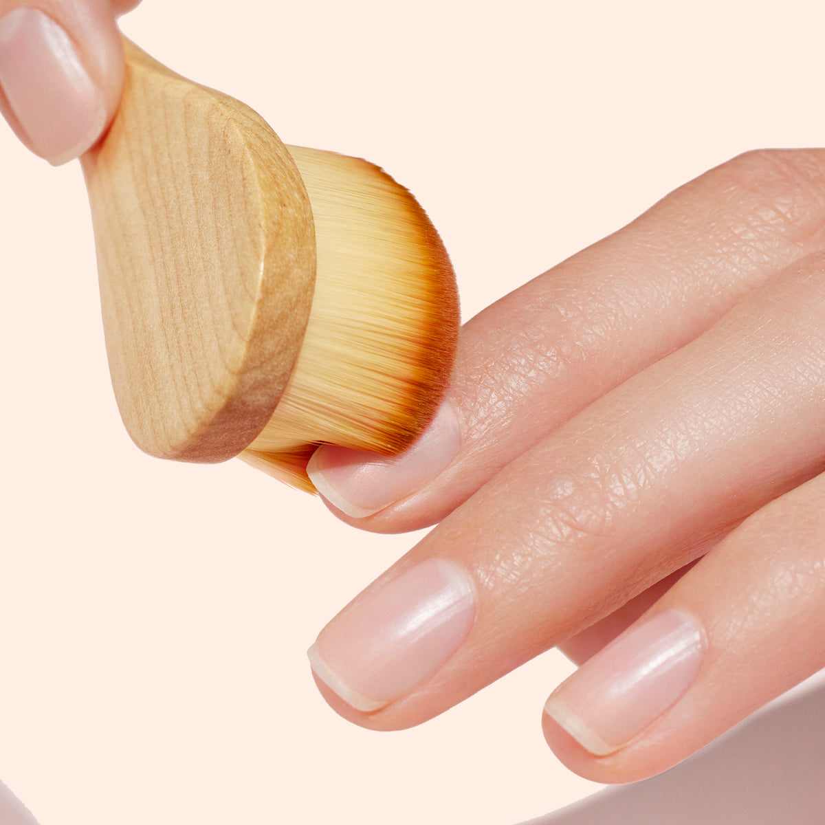 A close-up shows a hand with manicured nails being brushed by the Manucurist Dust Brush, made with vegan bristles and a wooden handle, gliding smoothly over the nails against a light beige background.