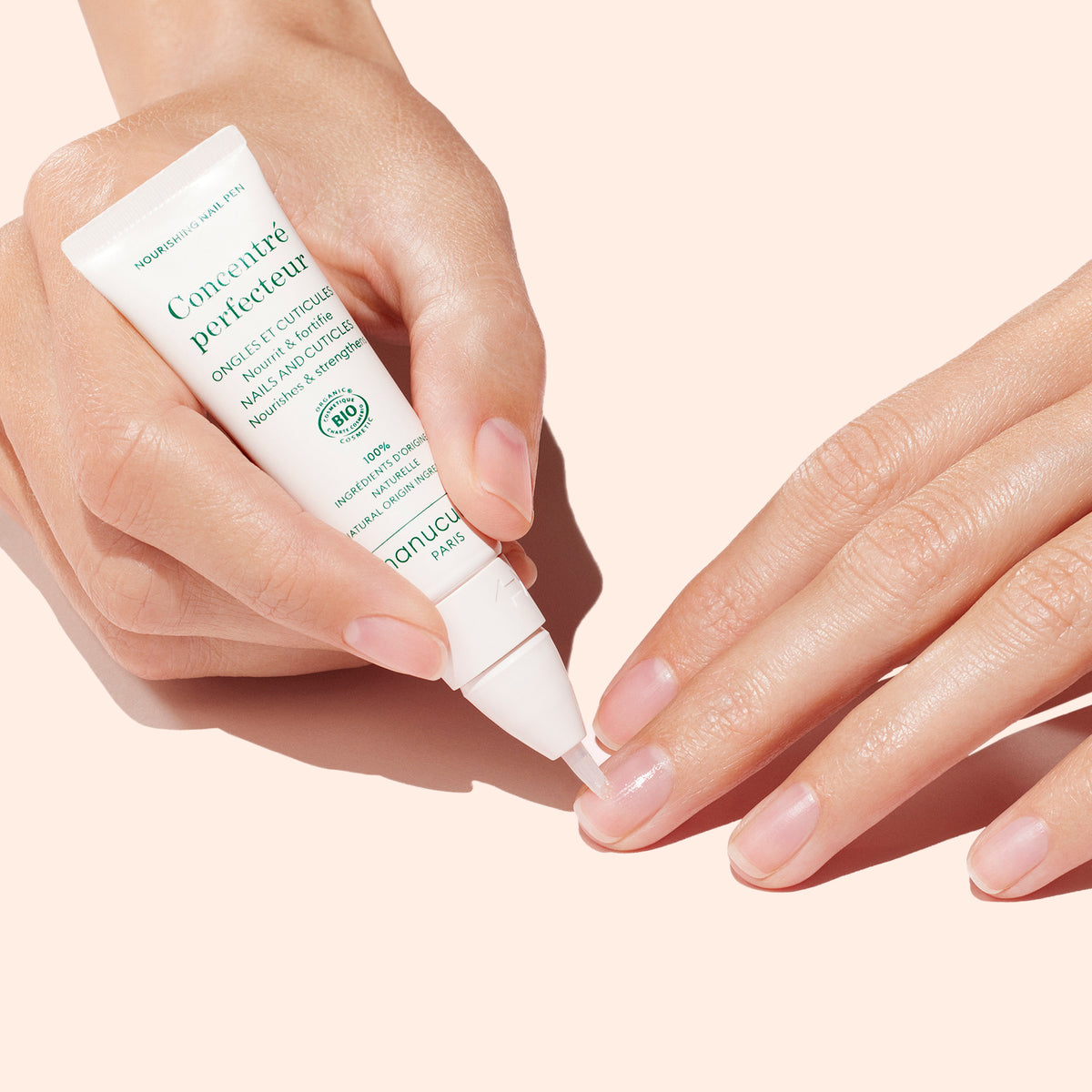 A hand applies Manucurist’s Nourishing Nail Pen - Cosmos Organic to the fingernails of the other hand for gentle cuticle care and stronger nails, set against a pale peach background.