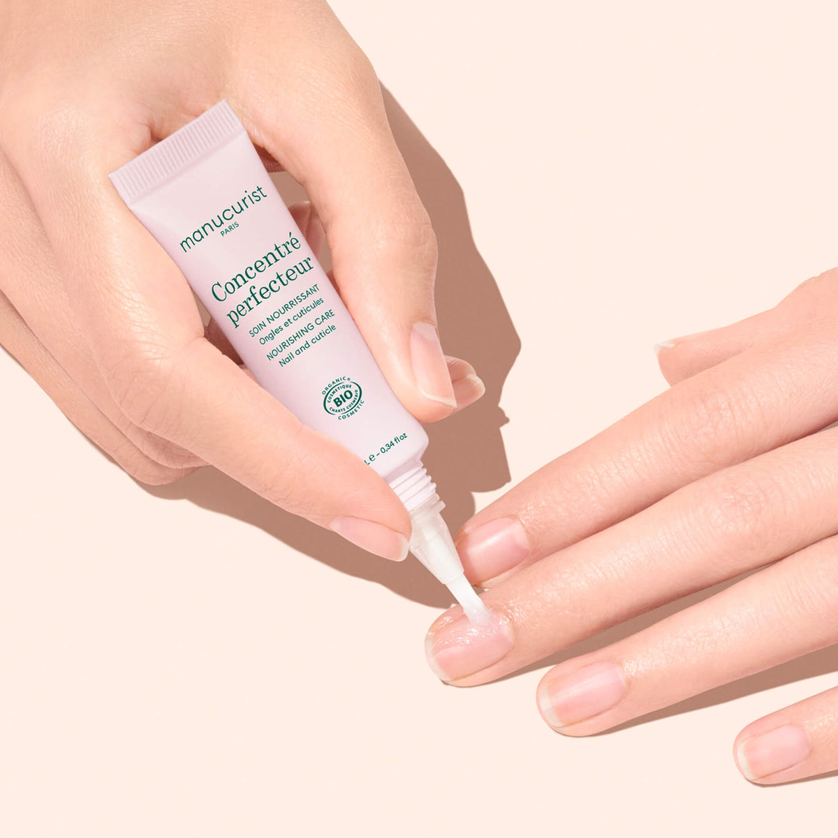 A hand applies a clear serum from a white tube labeled Manucurist Stronger Nails Duo onto the nails of the other hand, highlighting nail care against a light peach background.