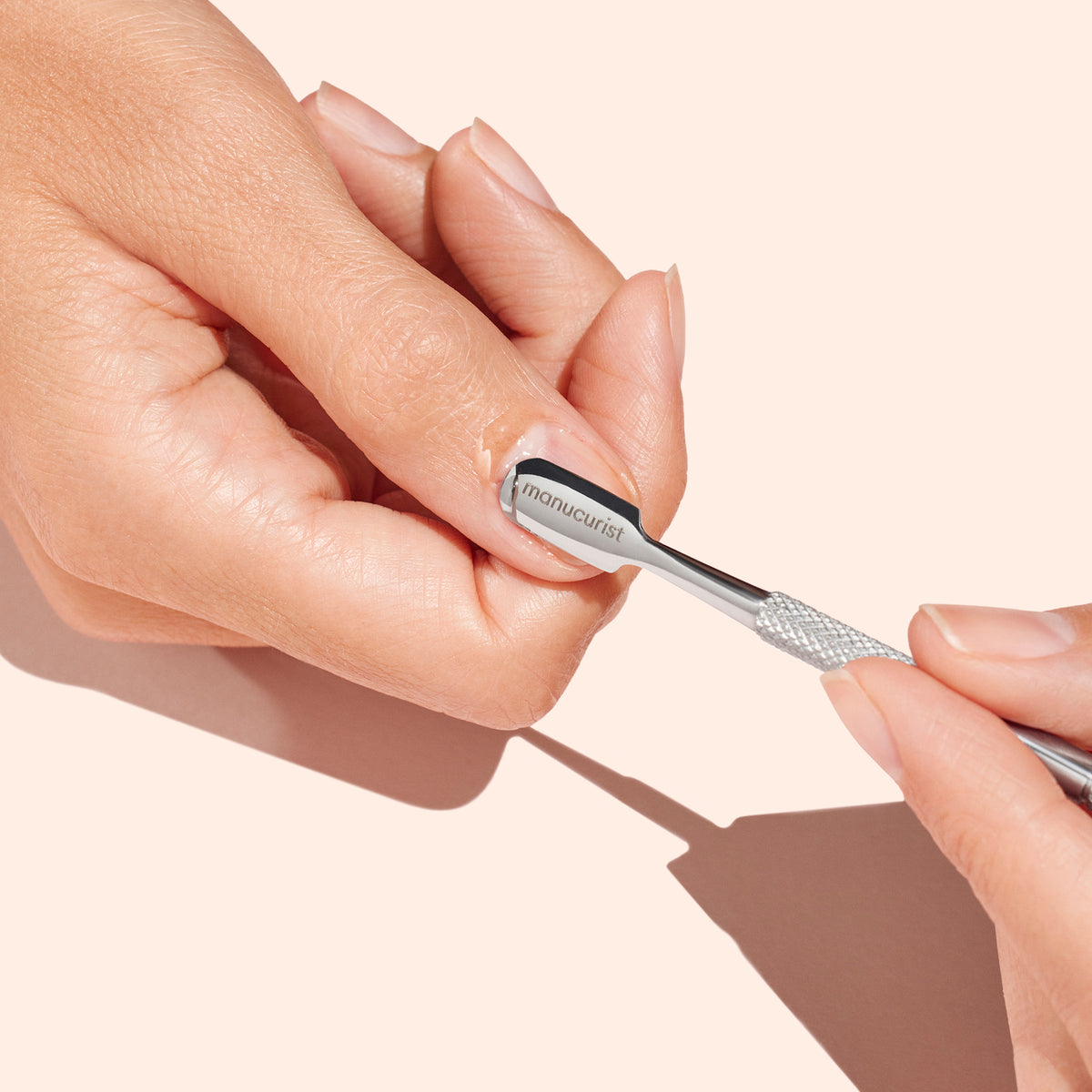A person uses the Manucurist Steel cuticle pusher to gently push back their thumbnail cuticles against a light beige background, showcasing careful nail care.