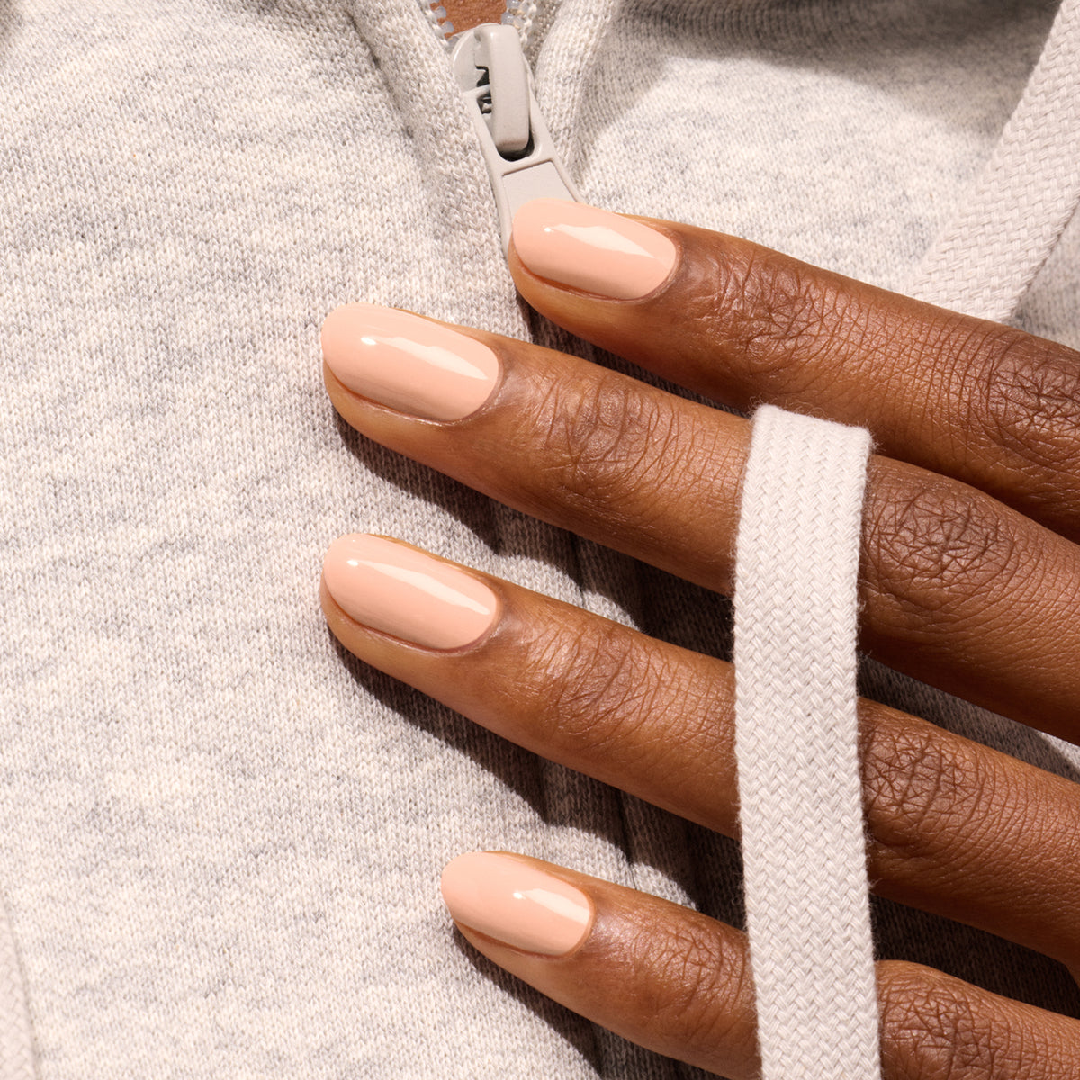 A hand with neatly manicured, glossy Pink Sand polish by Manucurist, a beige shade with subtle orange undertones, rests on a light gray zip-up hoodie, with one finger hooked around the white drawstring.