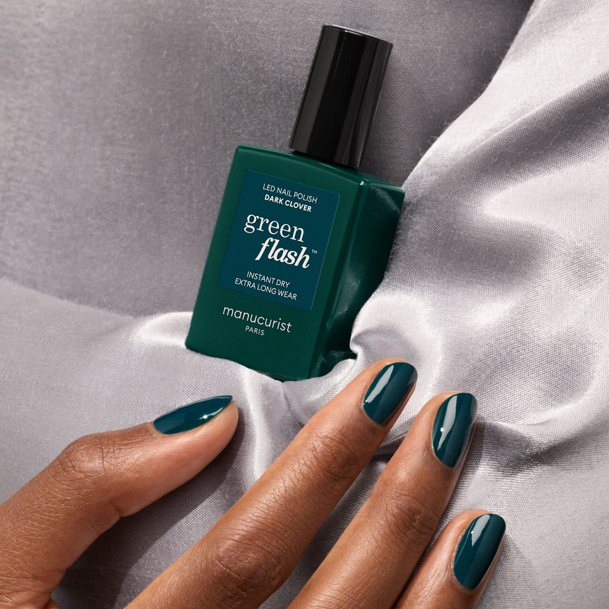A hand with glossy teal green gel polish rests on silver satin fabric beside a matching bottle of Dark Clover LED nail polish by Manucurist.