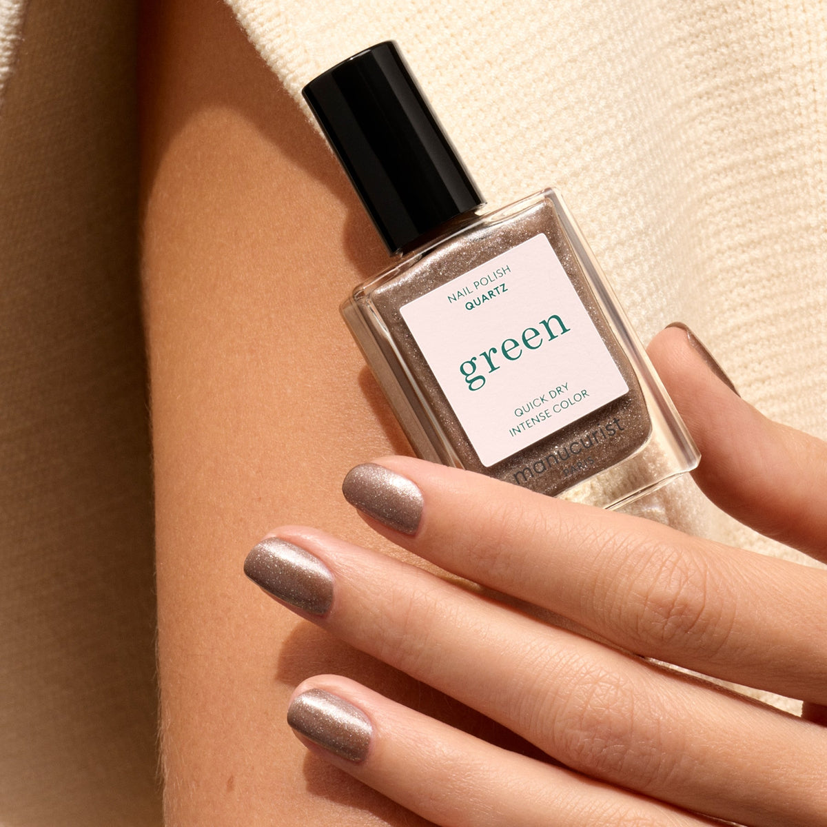 A hand with metallic taupe nail polish holds a square bottle of Manucurist Quartz nail polish with a black cap, set against beige fabric—pair it with frosted brown or sparkling silver glitter for extra flair.