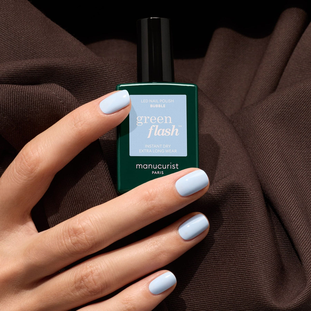 A hand with short, cool light blue manicured nails holds a green bottle of Manucurist Bubble nail polish against a dark brown fabric background.