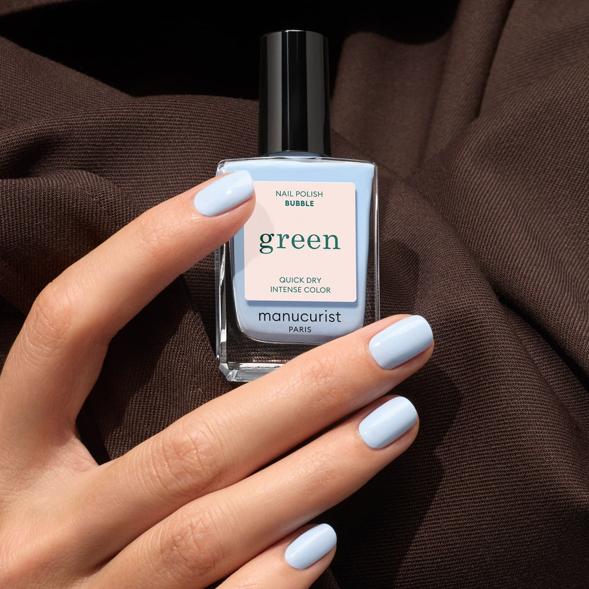 A hand with neatly manicured nails painted in Manucurist’s “Bubble” polish, a light blue shade with subtle grey undertones, holds the bottle from the brand’s Green™ range against brown fabric.