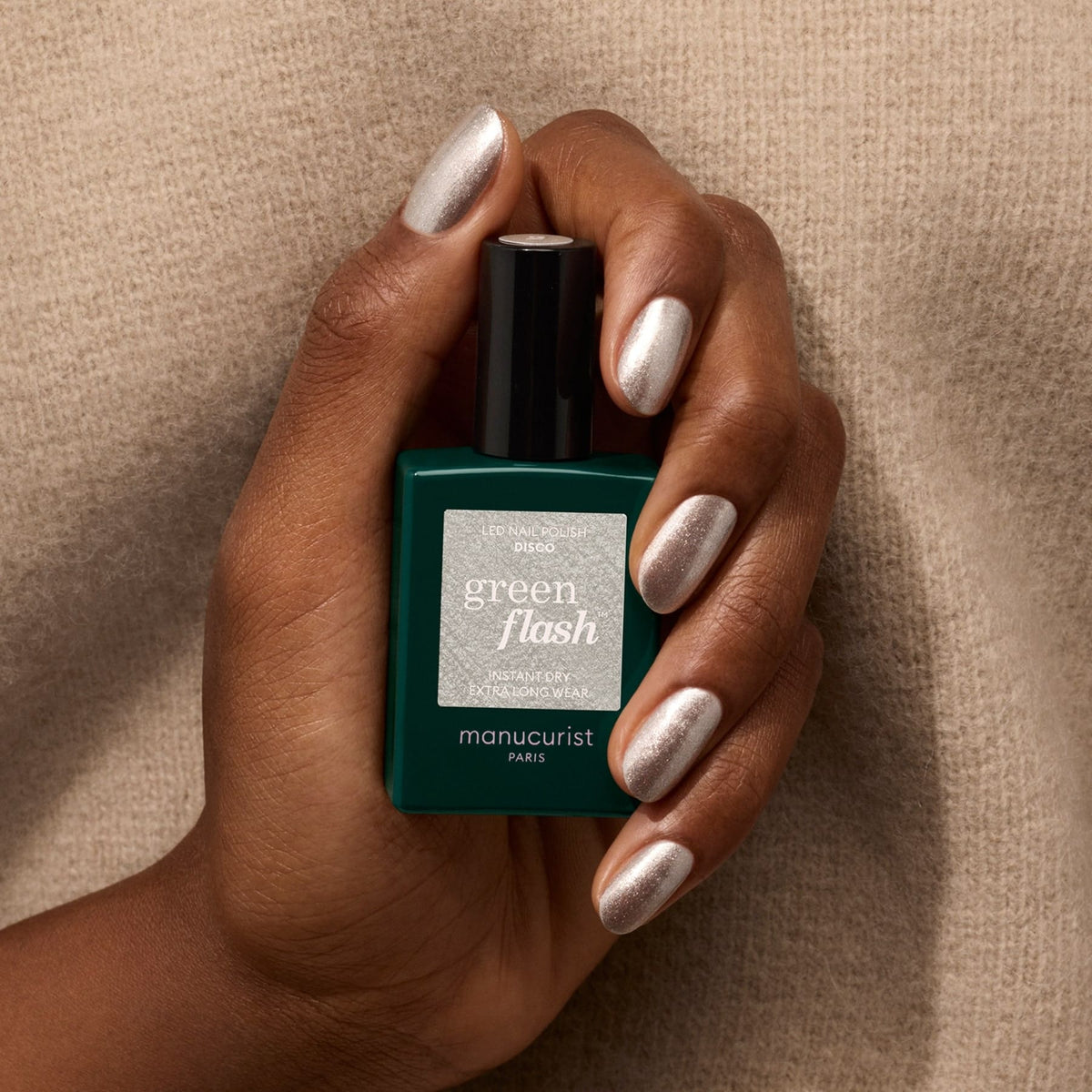 A hand with metallic, light pink nail polish holds a green bottle of Manucurist Disco polish against a beige textured background.