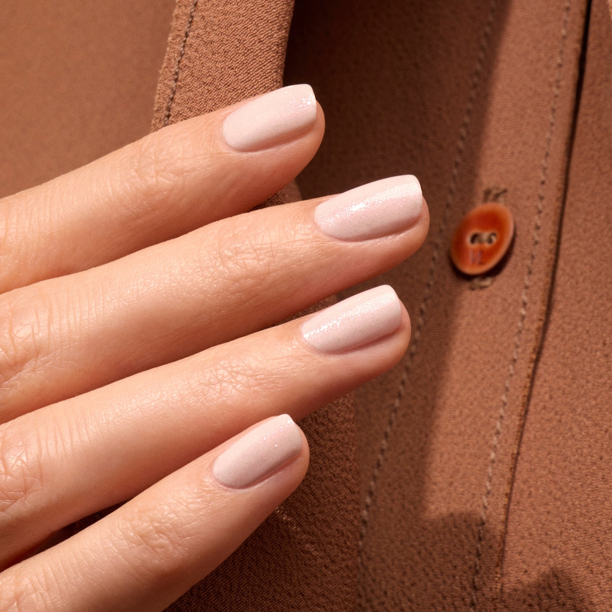 A hand with neatly manicured, iridescent pink shimmer nails, polished with Manucurist Gloss, rests against brown fabric with a button, highlighting the glossy finish and smooth skin.