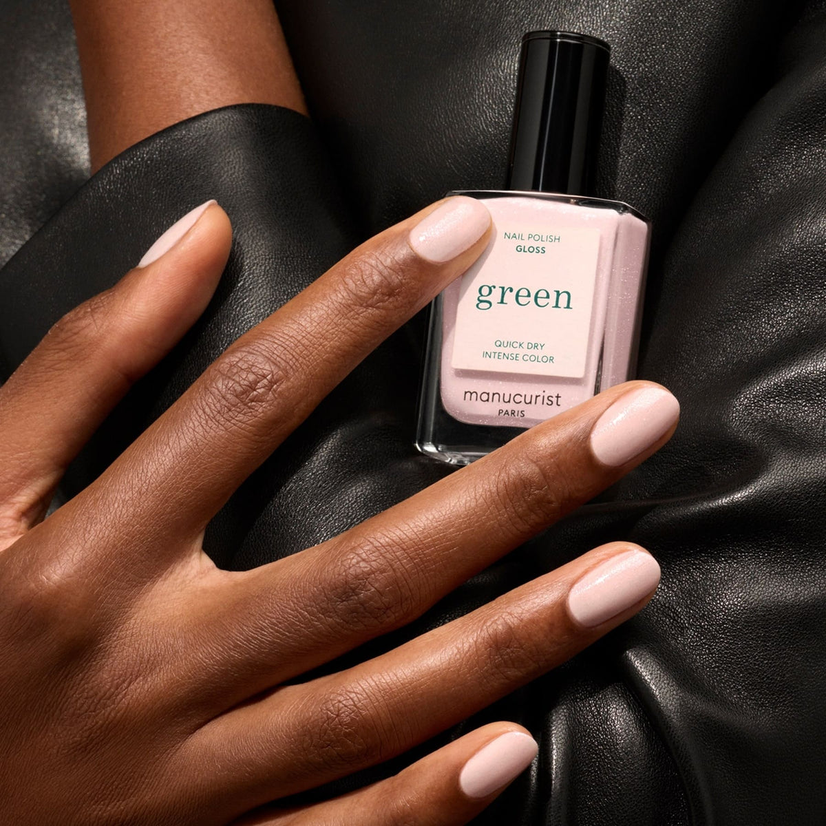A hand with iridescent pink shimmer nail polish rests on black leather, holding a bottle of Manucurist Gloss nail polish in a matching shade.