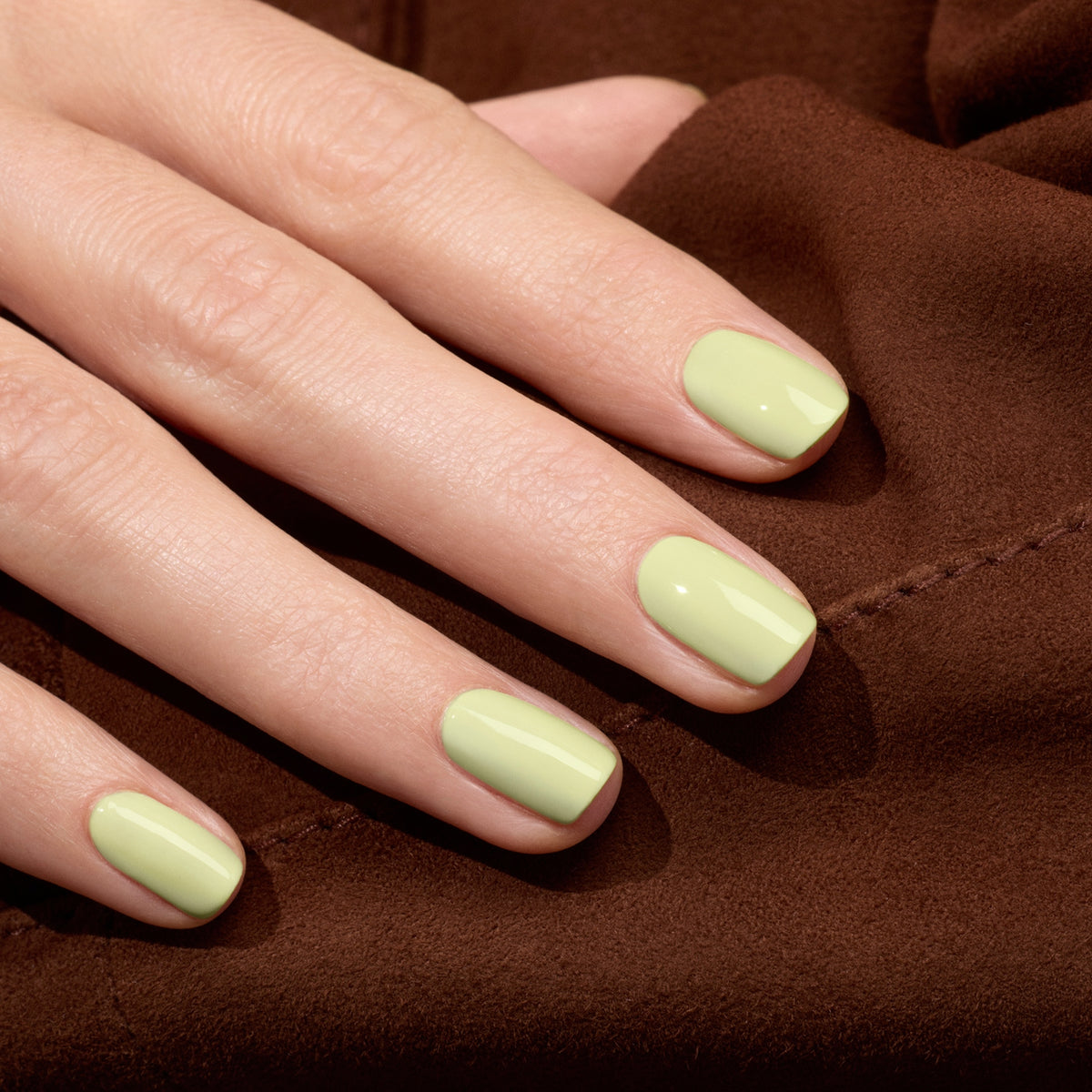 A close-up of a hand with neatly manicured nails painted in Manucurist Pistachio LED Gel polish, resting on a brown fabric surface.