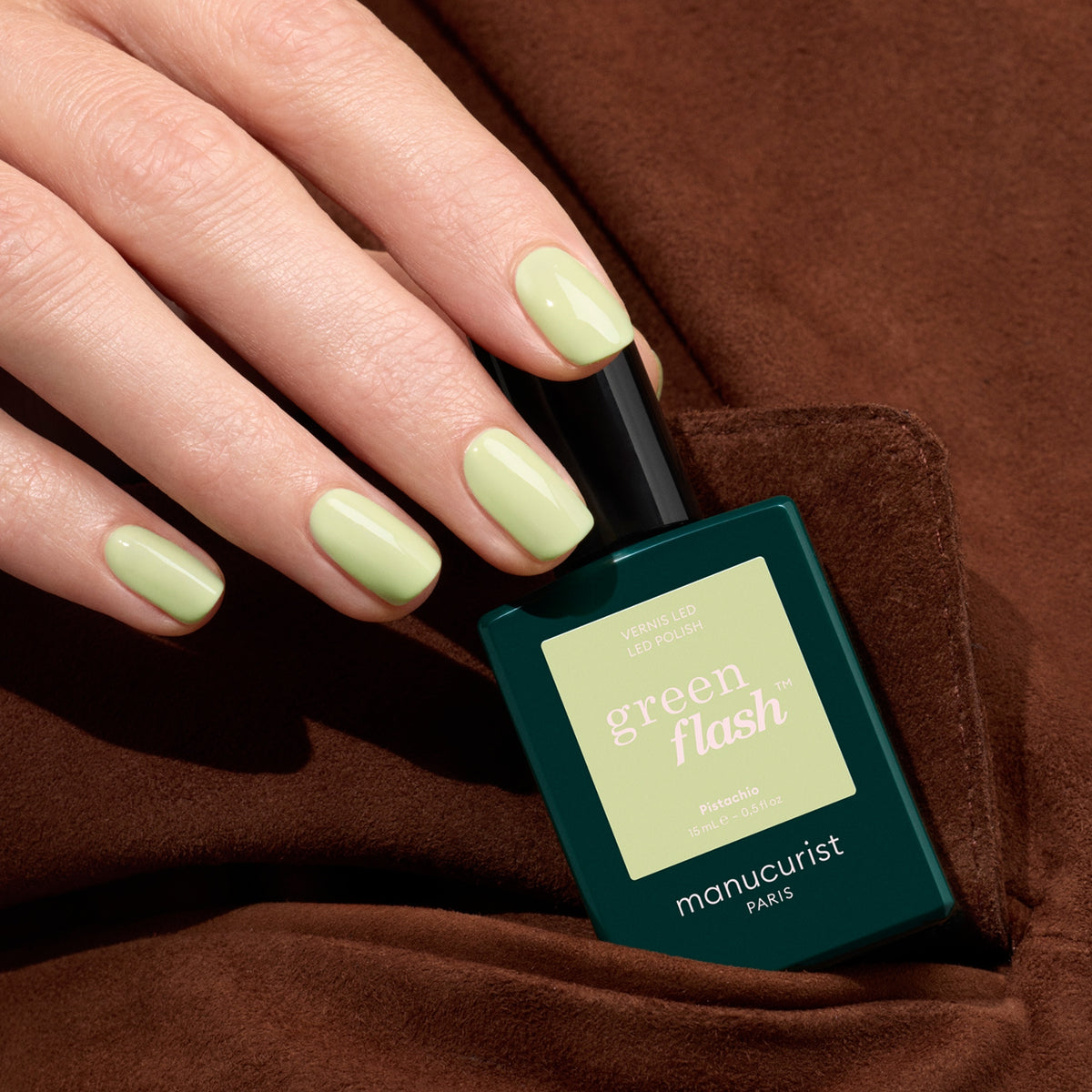 A hand with neatly manicured nails painted in pale green Pistachio polish from Manucurist holds the bottle against a brown fabric background.