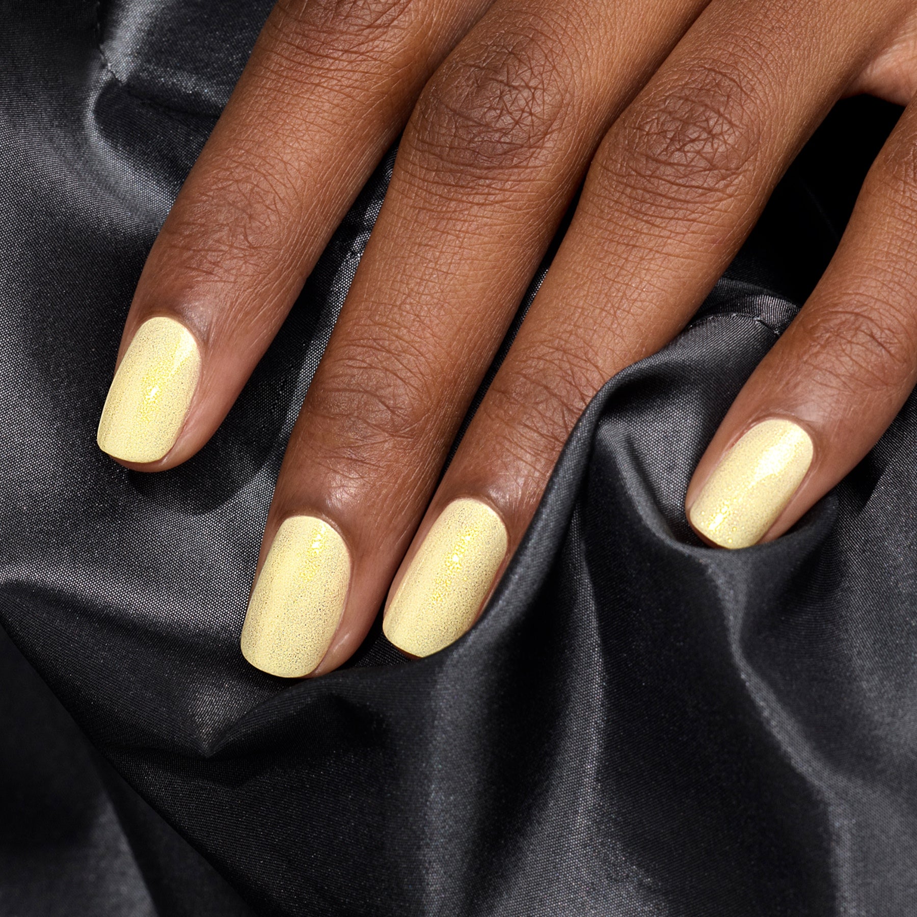 A hand with neatly manicured nails painted in Manucurist’s Lemonade, a pale yellow gel polish, rests on black satin fabric and showcases subtle glittering highlights.