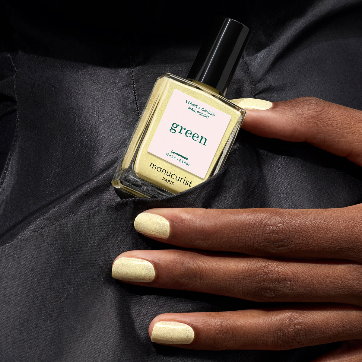 A hand with pale yellow polish holds a matching bottle of Manucurist Lemonade nail polish against a dark fabric background.