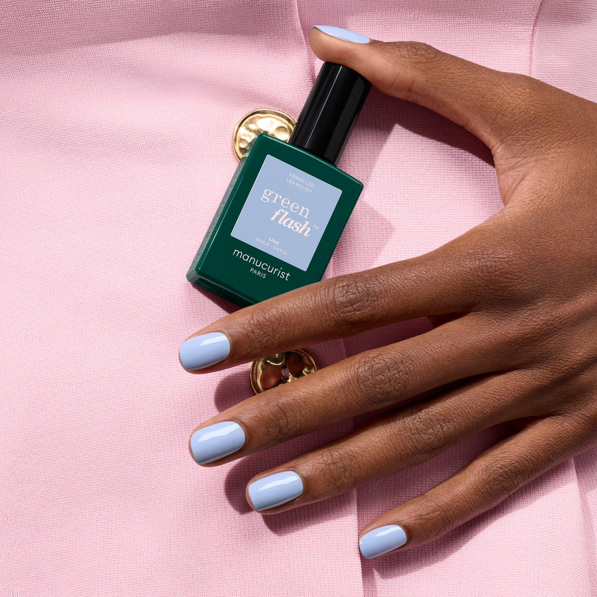 A hand with pastel blue gel polish holds a green bottle of Manucurist Lilas against a pale pink fabric background with a gold button.